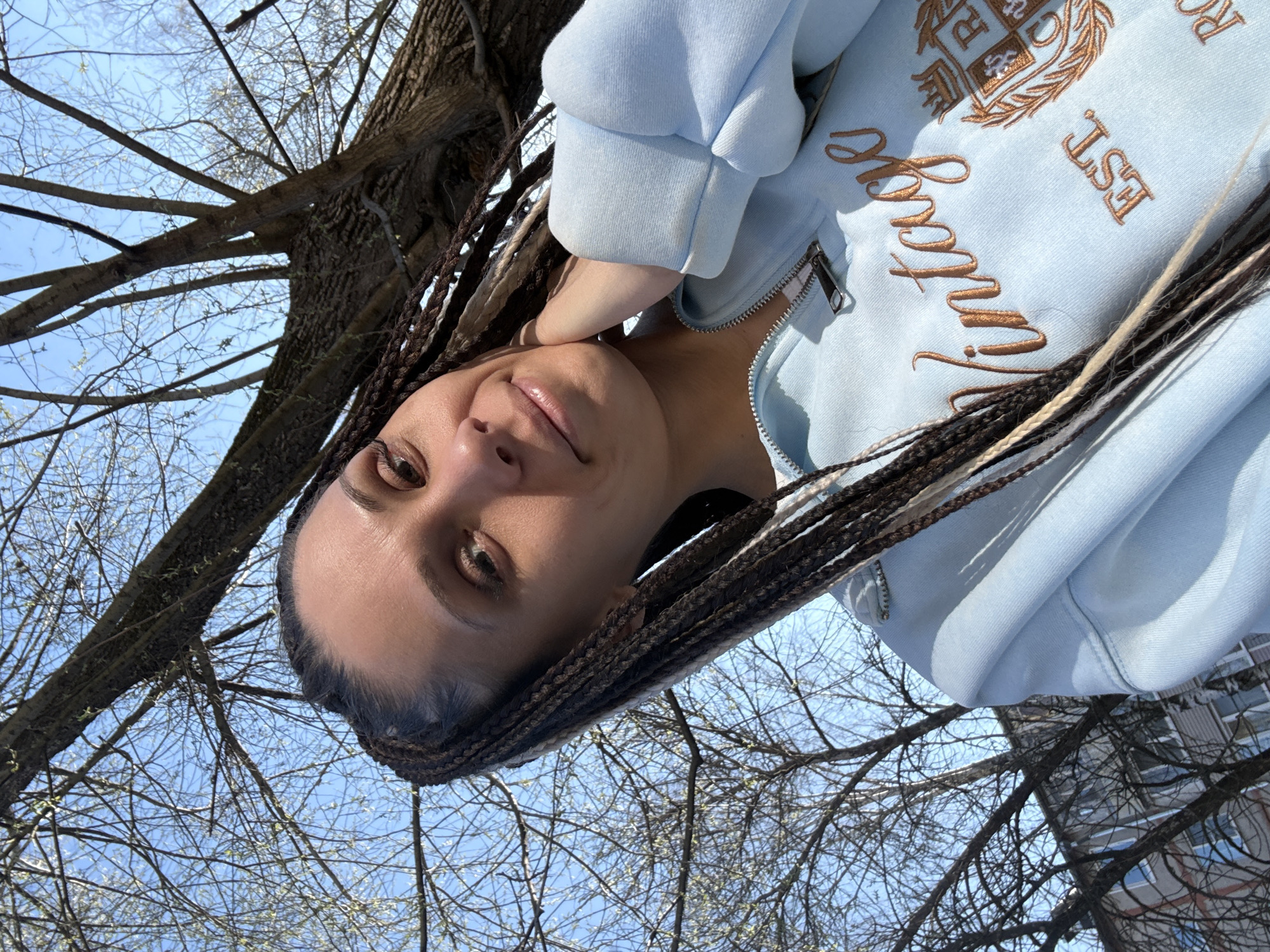 This media file is a rotated portrait of a young woman taken outdoors, appearing to be rotated 90 degrees clockwise. The woman's face, positioned on the left side of the frame, is looking upwards and slightly to her right, directly towards the camera, with a gentle, relaxed smile. She has dark eyes and long, dark braided hair interspersed with lighter, possibly blonde, strands that frame her face and extend towards the right.
She is wearing a light blue, zippered hoodie. The hoodie features prominent embroidery in a golden-brown thread on the right side of her chest. Visible text includes "Vintage" in a script font and "EST" in block letters. Above these, there is a crest-like design with initials (partially obscured, possibly "R" and "D" or "B") and decorative elements such as laurels. The zipper of the hoodie is also visible.
The background is dominated by a clear, bright blue sky, suggesting a sunny day. Numerous bare tree branches are silhouetted against the sky, indicating early spring or late autumn. Small, light green buds are discernible on some branches, hinting at the onset of spring. A thick tree trunk is visible in the upper right. Faintly, through the branches, a building with windows can be glimpsed in the distance.
The activity is a casual outdoor photo or selfie, captured from a low angle looking upwards. The overall atmosphere is pleasant and serene, consistent with a bright day in Rusanovo, Russia during spring.
