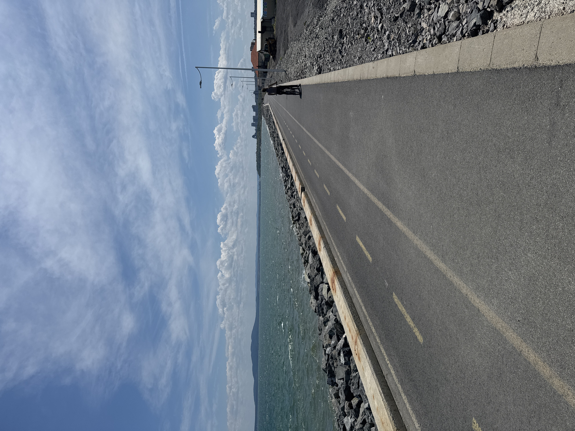 The image shows a paved path running alongside a body of water, likely the Black Sea, judging by the landscape. A person is riding a bicycle on the path. The path has yellow lane markings and a concrete edge bordering the water. A rock barrier separates the path from the water. On the other side of the path is a rocky area that grades into a building. Streetlights are visible in the distance along the path. The sky is blue with scattered clouds. Given the landscape and context, it is likely that this image was taken near Burgas, Bulgaria, along the coastal promenade.