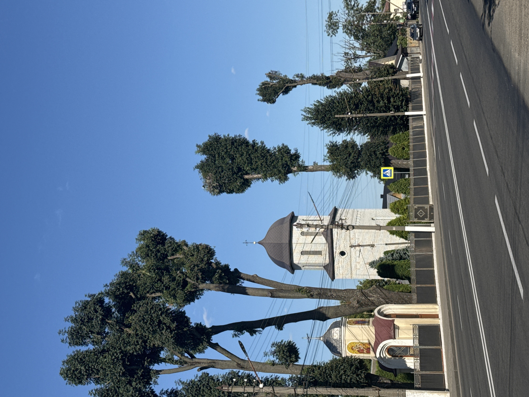 A street scene in an urban or suburban environment under a clear blue sky. On the left side of the road, there is a small church with a dark dome and light-colored brick construction, adorned with religious icons above its entrance. Adjacent to it is a taller bell tower, also constructed of light-colored bricks. A dark metal fence runs along the property line, separating it from the road. Several tall trees with dense green foliage are situated behind the church and tower, with one tree on the right appearing to have been pruned into a distinct shape. Across the street, houses with wooden roofs and a black metal fence are visible. A pedestrian crossing sign is positioned near the houses. Two cars are parked on the right side of the road, and a third is driving away in the distance. The road itself is asphalt with double white lines marking the lanes. Power lines are visible strung between poles throughout the scene.