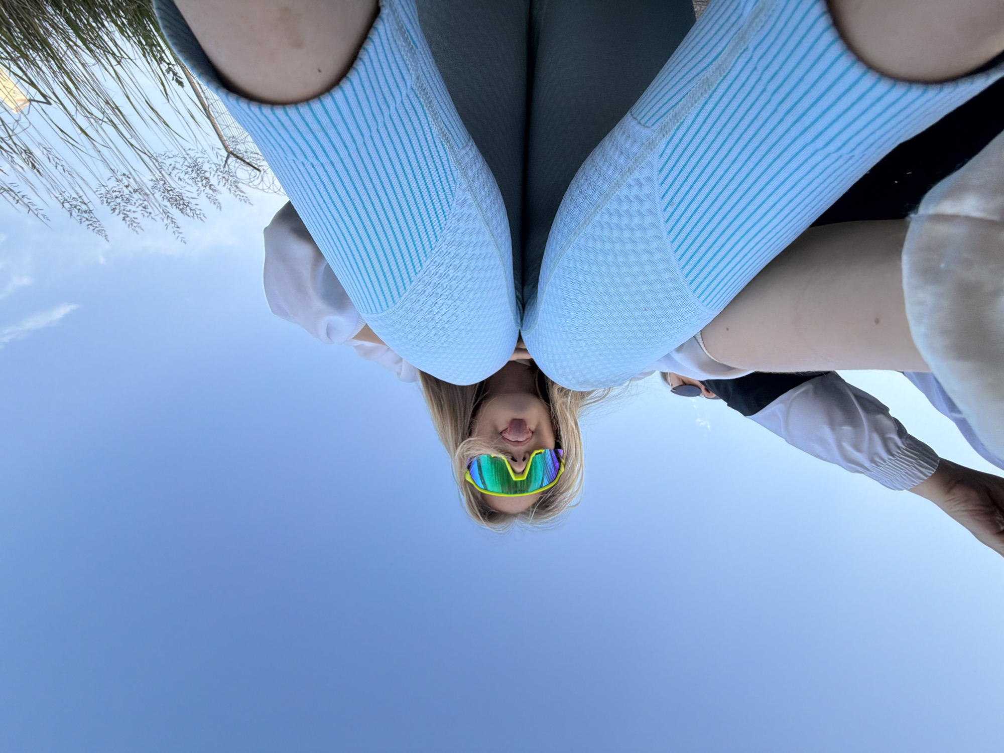 An outdoor, low-angle, wide shot shows a young woman with blonde hair wearing neon green and blue ski goggles and sticking her tongue out. She is wearing a white jacket over a black shirt and light blue leggings. The background is a clear blue sky with a few wisps of white clouds and some dry grass on the right side of the frame. The image appears to be taken in Chaikovsky, Russia, during daylight hours.