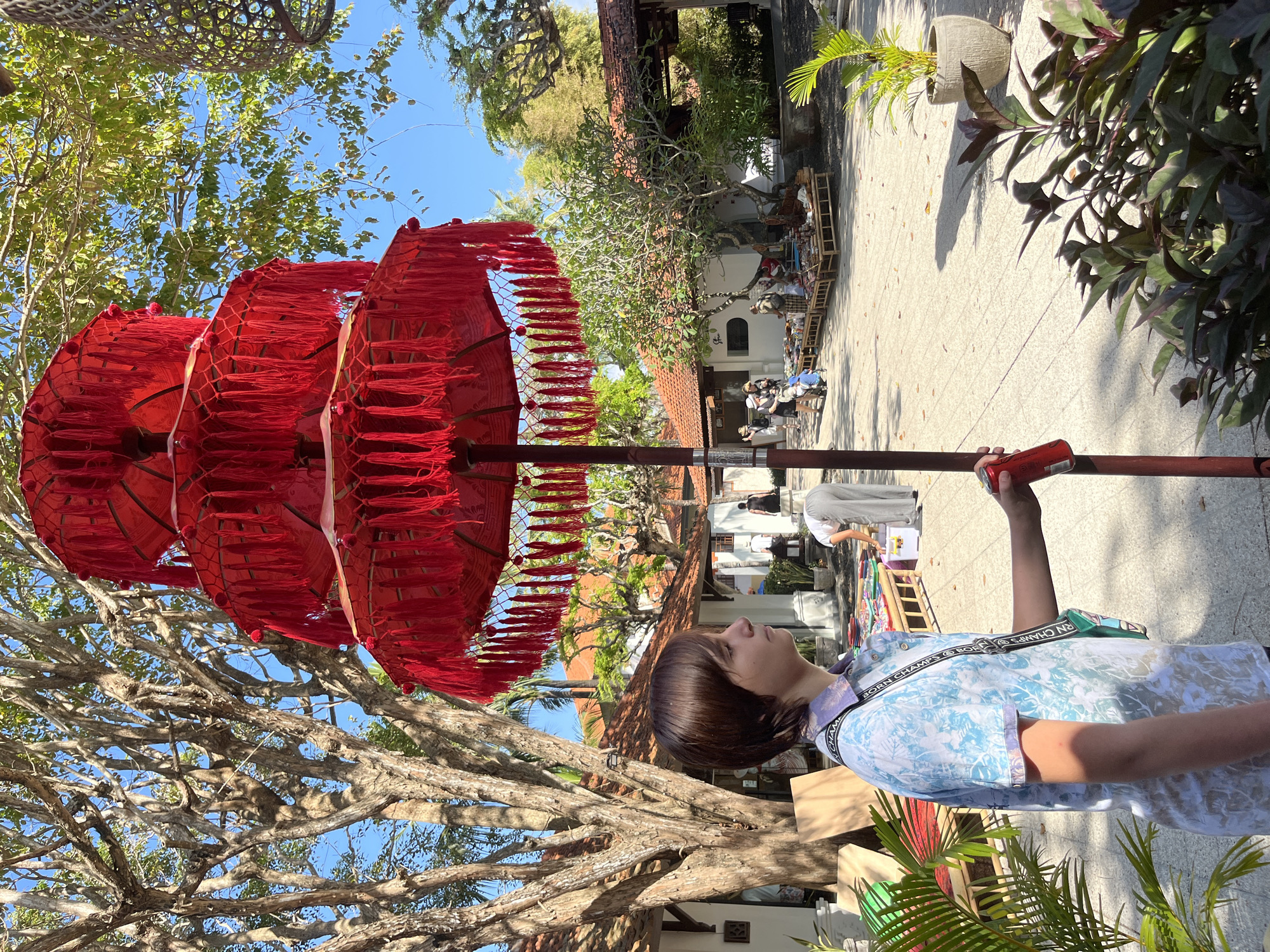 In Kuta Selatan, Indonesia, a young person stands in a courtyard with a red, tiered umbrella. The umbrella is decorated with fringes and sits atop a pole. The courtyard is paved and has buildings with terracotta roofs. In the background, a market stall displays various items for sale. Trees with intricate branches and lush green foliage are scattered throughout the scene, and the sky is clear and blue. The young person is wearing a patterned shirt and a crossbody bag and is holding a red can. The overall atmosphere is bright and sunny, suggesting a warm day.