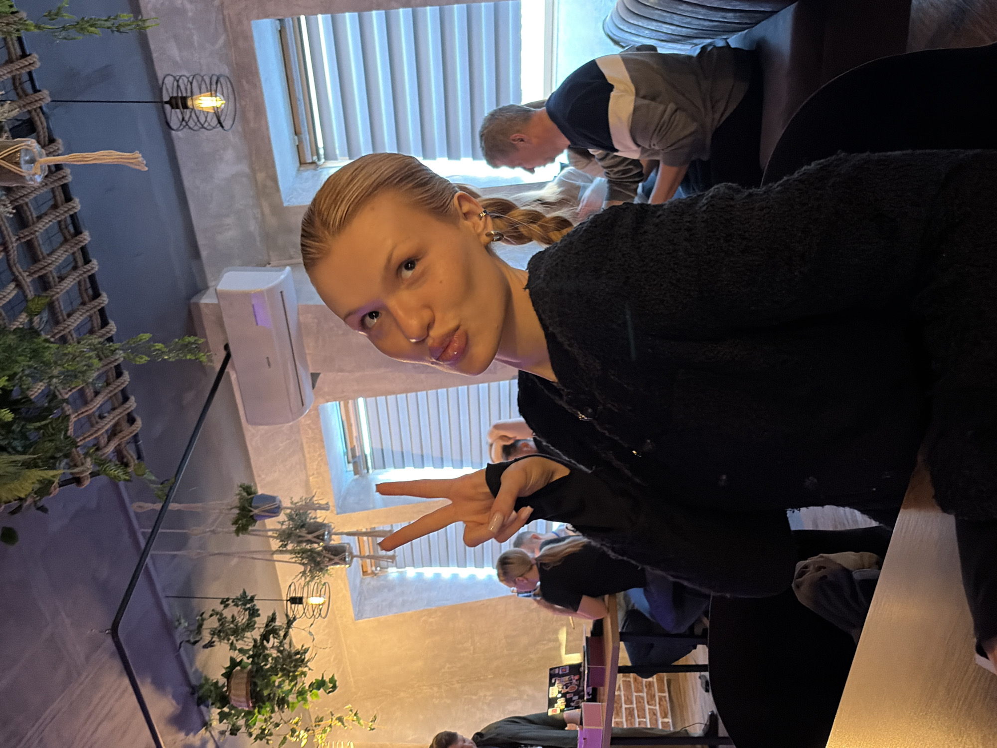 An indoor scene in a cafe or restaurant located in Chaikovsky, Russia, features a young light-skinned female in the foreground, seated at a dark wooden table. She is wearing a dark, textured sweater, has blonde hair pulled back, and is looking directly at the camera, pouting her lips while making a peace sign with her right hand. Her left ear is visible, adorned with multiple earrings.
In the mid-ground and background, several other individuals are present. To the left, a male figure in a dark jacket is partially visible near a second dark wooden table. On this table, a laptop displays colorful elements, and several pink boxes are present. A female with blonde hair in a ponytail and a black shirt is seated at the same table, looking at a smartphone. To the right, a male with short reddish-brown hair, wearing a horizontally striped sweater (white, grey, and dark blue), is seated on a dark brown upholstered bench, his profile turned towards the left. Another partially obscured female is seated next to him.
The environment features light-colored, textured walls, with a section of exposed brickwork near the floor on the left. Multiple windows fitted with horizontal blinds emit a cool blue light. The dark ceiling is decorated with artificial hanging plants, macrame plant hangers, a rope lattice structure, and industrial-style pendant lights with exposed Edison bulbs. An air conditioning unit is mounted on the wall to the right. The overall lighting is a mix of warm artificial light and the cool ambient light from the windows.