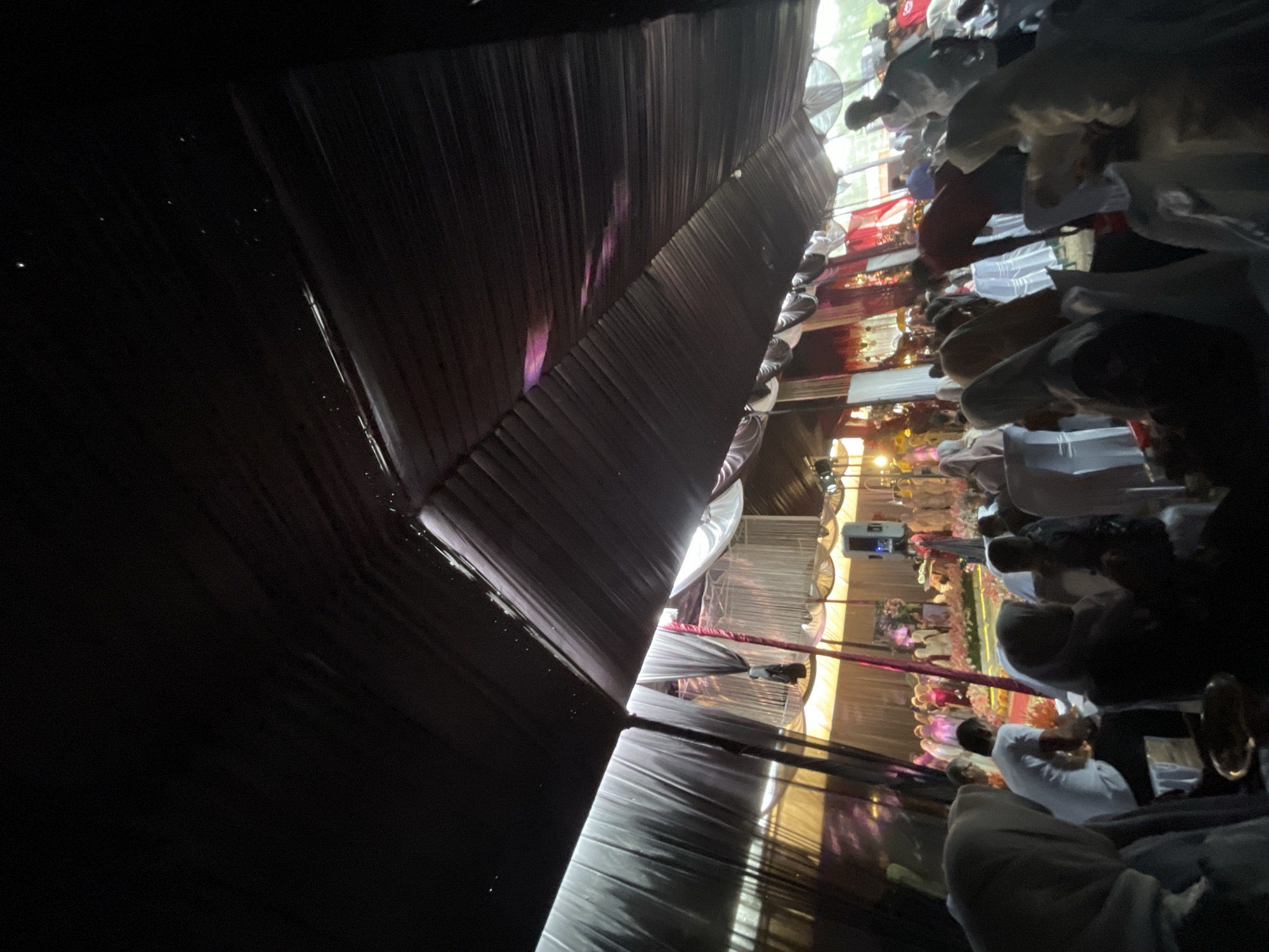 The image depicts an indoor event, possibly a wedding or a religious gathering, taking place under a large tent structure in Bulohlhoksukon Timur, Indonesia. The tent has a dark, draped ceiling and is supported by poles. The lighting is dim, with some areas illuminated by warm, yellow and pinkish light, suggesting artificial lighting and possibly decorative elements.
In the foreground, several individuals are visible, mostly wearing headscarves, indicating a likely cultural or religious context. They are seated, facing towards a central area where the event is unfolding. The atmosphere appears to be one of communal gathering and observation.
The midground and background reveal more attendees and the main focal point of the event. There is a decorated stage or platform area with floral arrangements. People are standing and seated around this central area, some dressed in formal attire. The overall scene suggests a vibrant and formal social occasion within a community setting.
