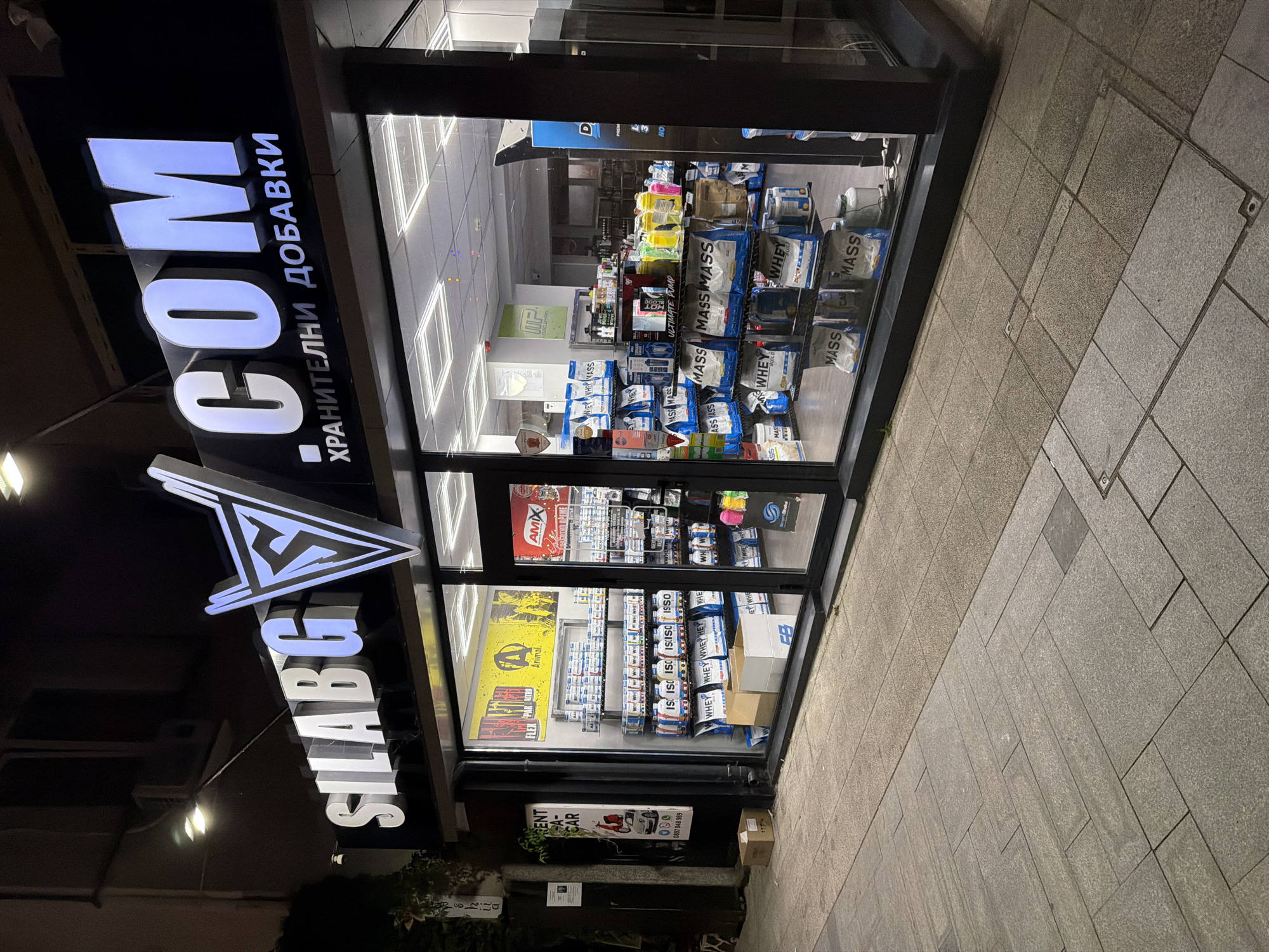 This is a nighttime, eye-level, exterior shot of a storefront for "SILABG.COM", a store specializing in nutritional supplements, located on Bogoridi Street in Burgas, Bulgaria. The facade features a large, illuminated sign with the store name. The storefront itself is a large glass window displaying rows of various supplement products, including bags of protein powder and tubs of vitamins. The interior is brightly lit, with fluorescent ceiling lights. The sidewalk in front of the store is paved with grey and brown tiles, and there are a few cardboard boxes on the ground near the entrance. The street is dimly lit, with some ambient light from the store's signage and interior. A portion of a building is visible to the left of the storefront, with an air conditioning unit and some foliage.