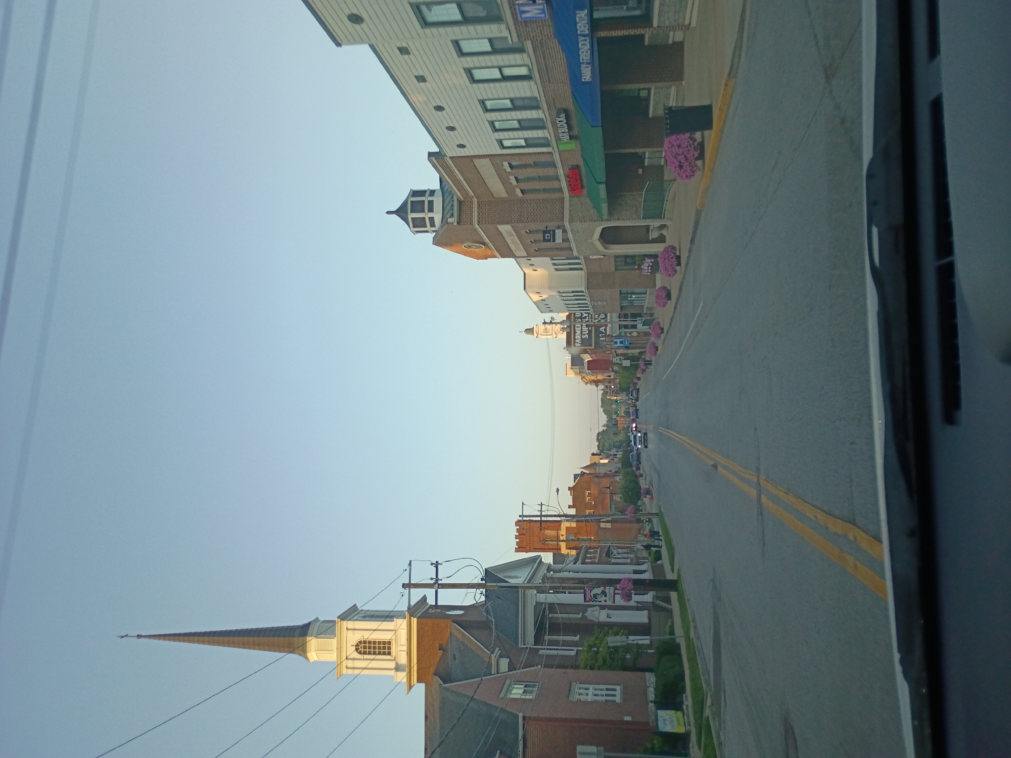 The image captures a view of a street in Versailles, United States of America, likely in the early morning or late afternoon given the soft lighting and clear sky. The perspective is from inside a vehicle, looking forward through the windshield and dashboard.
On the left side of the street, a prominent brick church with a tall white steeple and cross is visible. Adjacent to the church is a building with a white facade and columns. Further down the street on the left, there are more brick buildings and trees.
The right side of the street features a row of commercial buildings. These buildings are a mix of brick and lighter-colored facades, with awnings and storefronts. Signs indicate businesses such as "FARMERS U SUPPLY", "KICKBACK", "HAIR BLOCK", and "FAMILY FRIENDLY DENTAL". Potted pink flowers line the sidewalk in front of these businesses.
The street itself is paved and has double yellow lines in the center. Several cars are driving on the street, and a few pedestrians can be seen walking on the sidewalk. The overall scene depicts a quiet, pleasant street in a small town.