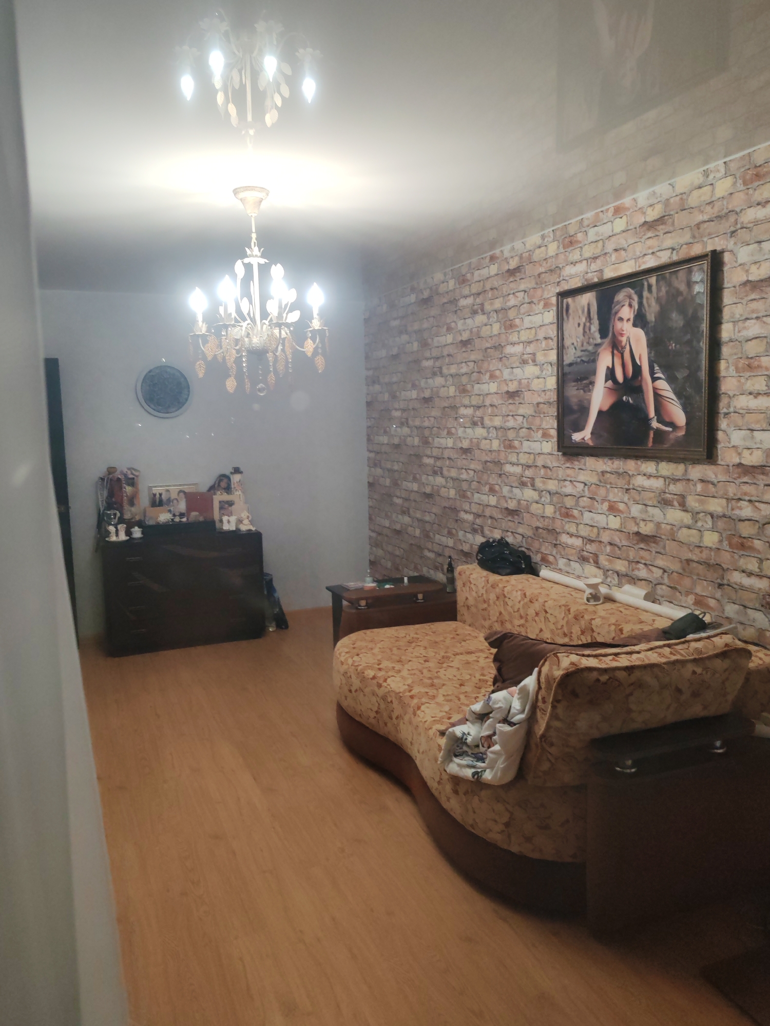 The image shows a room interior with a brick-patterned wall on the right. On this wall is a framed portrait of a woman. In the center of the room is a floral patterned sofa. To the left of the sofa is a small dark-colored side table. A dark-colored dresser is located against the left wall, covered with decorative items and pictures. Two chandeliers hang from the ceiling. A partial view of a white curtain is seen on the left edge of the image. The room has wood-look flooring.
While the image provides an interior view, there is no discernible information in the image to determine the location as Petropavlovsk-Kamchatsky, Russia.