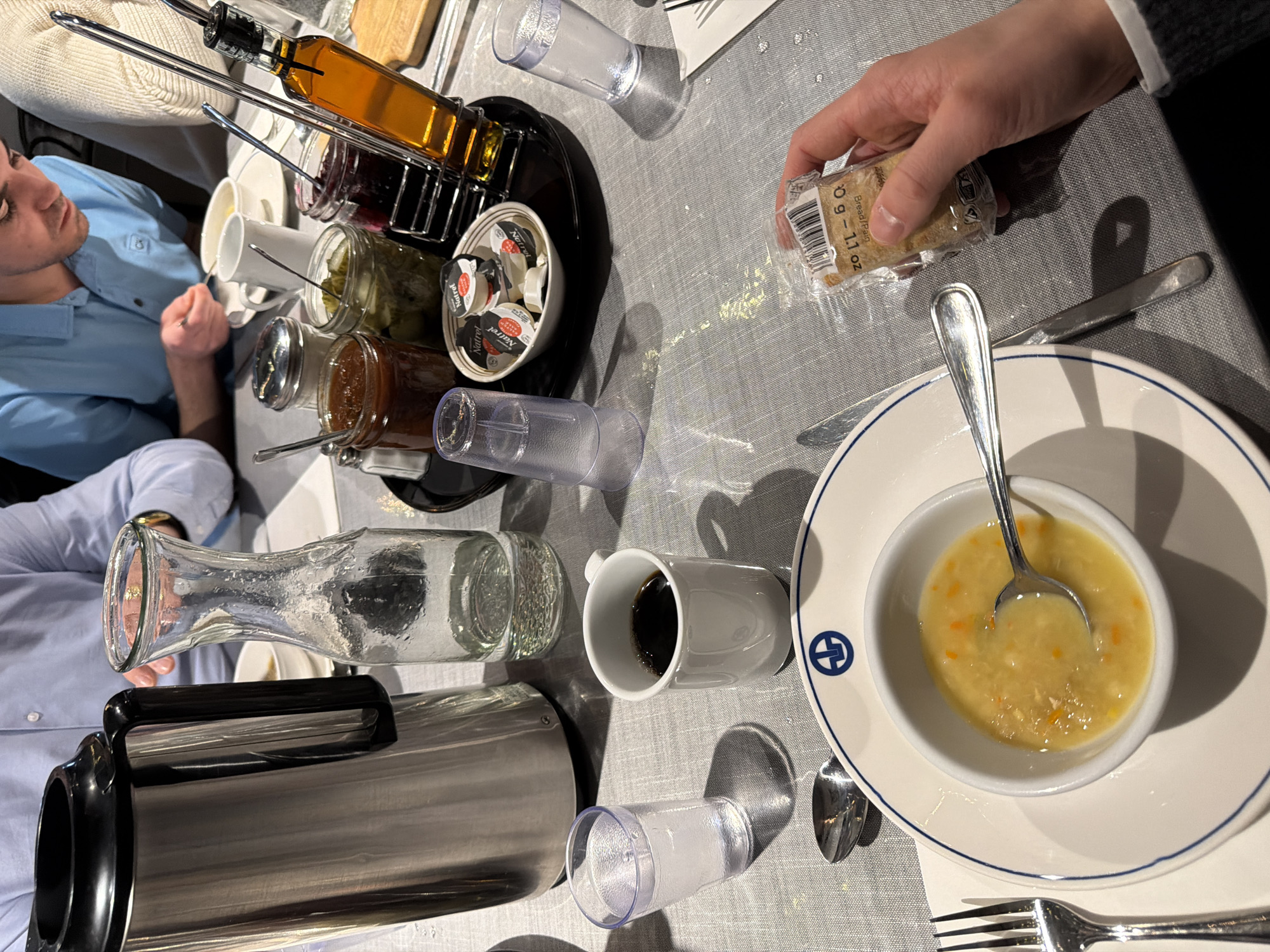 A top-down perspective captures a breakfast scene. A man in a blue shirt sits to the left. Various breakfast items are arrayed across the table. There's a rack holding multiple jars of condiments, including what appears to be honey, jam, and pickles. A bowl containing individual creamers sits nearby.
A person's hand on the right holds a small package of bread. A bowl of soup with a spoon sits on a plate in the lower right. A coffee mug filled with dark liquid and a glass water carafe are also present. A metal coffee server sits on the bottom left. Cutlery is arranged around the soup bowl. The location is unknown and cannot be ascertained from this image. The city of Mirabel, Canada is also unable to be discerned from this image.