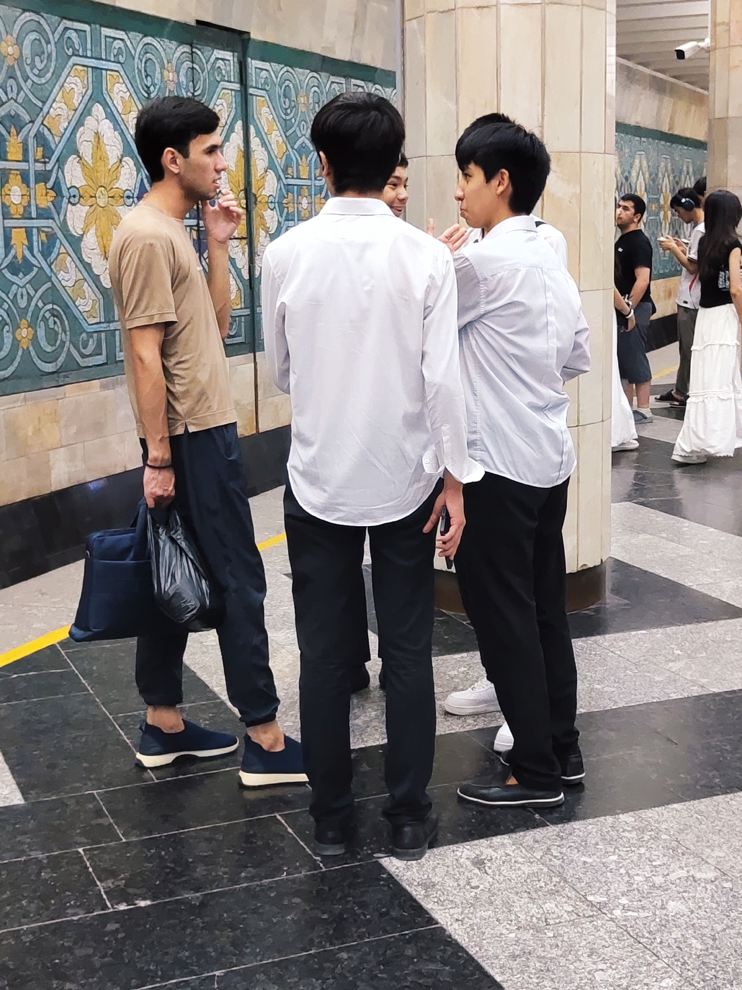 The image depicts a group of young men standing in what appears to be a public transport station or a large indoor public space, likely in Tashkent, Uzbekistan, given the distinctive architectural and decorative elements visible. The background features a wall with ornate, colorful tiles in a geometric pattern, characteristic of Central Asian design. The floor is a polished black and white checkered tile pattern.
In the foreground, three young men are the primary subjects. The man on the left, wearing a tan t-shirt and dark blue trousers, holds a dark blue bag and a black plastic bag in his left hand. He is looking to his right, with his right hand near his mouth, as if in conversation or deep in thought.
Directly in front of him is another young man wearing a white button-down shirt and black trousers. He is facing away from the camera, partially obscuring the view of a third young man to his right. This third man is also wearing a white button-down shirt and black trousers and is turned slightly to his left, facing the man in the tan t-shirt.
Further in the background, other individuals are visible, including a man in a black t-shirt and dark shorts, and a woman in a white long skirt and dark top, suggesting this is a busy public area. The overall impression is of a casual gathering or waiting area. The lighting appears to be artificial, typical of an indoor environment.