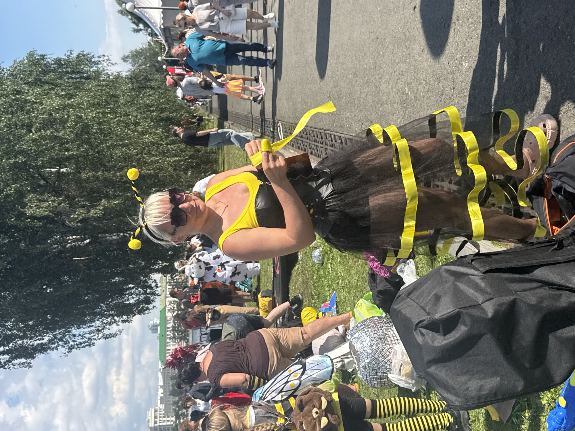 The image depicts an outdoor event in Yekaterinburg, Russia, likely a festival or parade, given the costumes. In the foreground, a woman dressed as a bee, with black and yellow attire and antennae, is the central subject. She is holding a roll of yellow ribbon. Behind her, other individuals are visible, some in costumes, including what appears to be a cow costume. Various items, such as bags, a disco ball, and a water gun, are scattered on the grassy area. In the background, trees line a pathway where people are walking, and buildings can be seen in the distance, suggesting an urban setting. The sky is partly cloudy, and the sun is shining. The overall scene suggests a festive and public gathering.