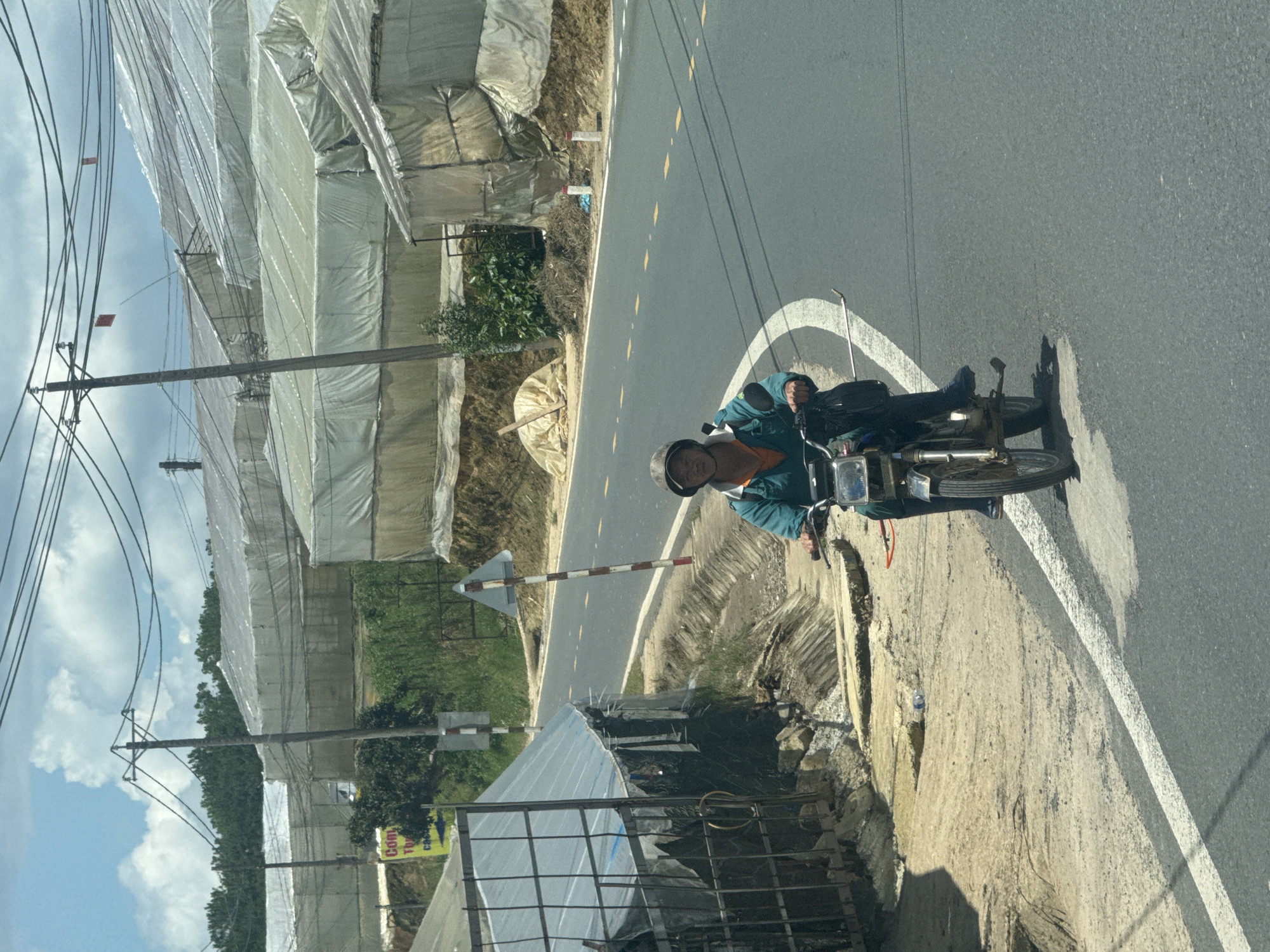 A person wearing a green jacket and a helmet is riding a motorcycle on a paved road in what appears to be a rural or semi-rural area. The person is looking towards the camera. The road has yellow dashed lines indicating lanes. On the left side of the road, there is a structure with a metal fence and a translucent roof, and further back, a hillside with greenhouses covered in white plastic. On the right side of the road, more greenhouses are visible, as well as power lines and utility poles. The sky is partly cloudy. A yellow sign with red text is visible on the left side, partially obscured by trees.