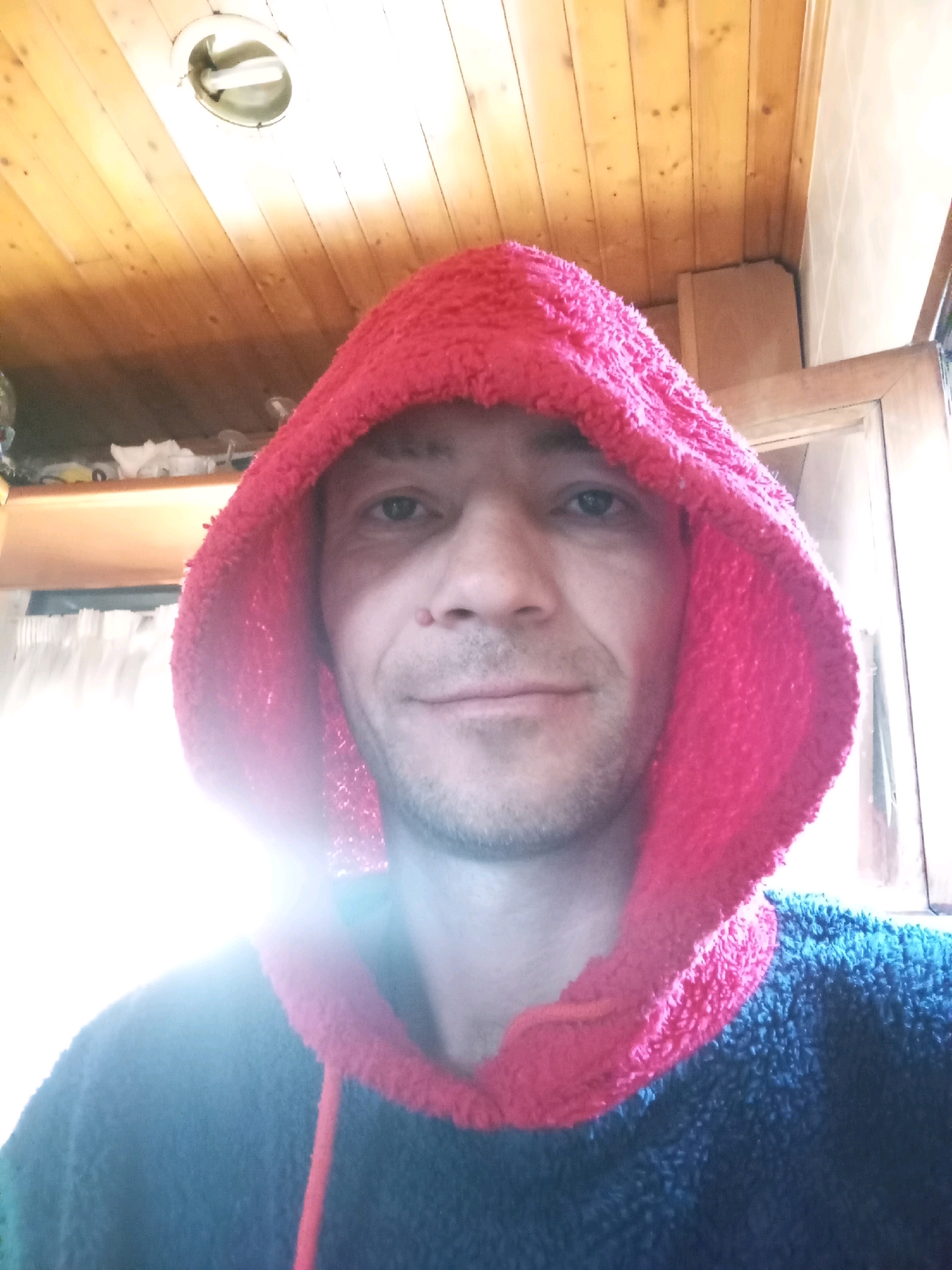 The image is a close-up, selfie-style photograph of a man wearing a red hooded sweatshirt. The man is looking directly at the camera, with a neutral expression. The background is dimly lit and appears to be an indoor setting, possibly a room with wooden paneling on the ceiling. The lighting in the image is somewhat harsh, with a strong light source coming from the upper left, causing lens flare. There are no discernible landmarks or indicators that would place this image in the city of Viseu, Portugal.