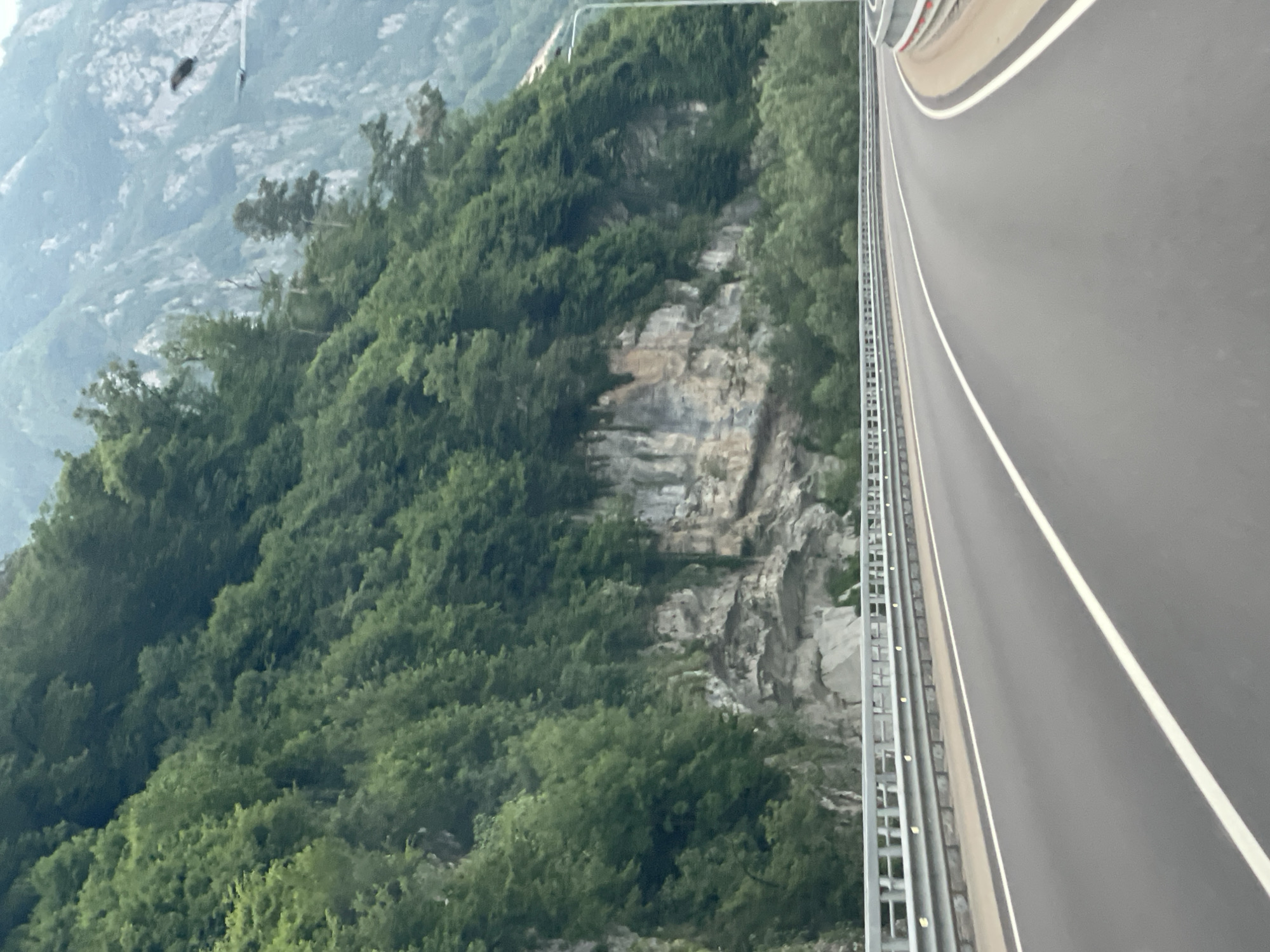 The image depicts a curving asphalt road with white lane markings, bordered by a metal guardrail on the left and a sandy shoulder with a red-and-white barrier on the right. To the left of the road, a steep, tree-covered mountainside rises dramatically, with exposed rock faces visible in sections. In the distance, further mountains are shrouded in a hazy, overcast sky. A streetlamp is visible on the right side of the road, positioned on the mountainside. No people are present in the image. The scene suggests a natural, mountainous environment, likely a highway passing through a scenic area. The description is not specific to the city of Monastyr, Russia, as no identifiable landmarks or contextual clues place it there.