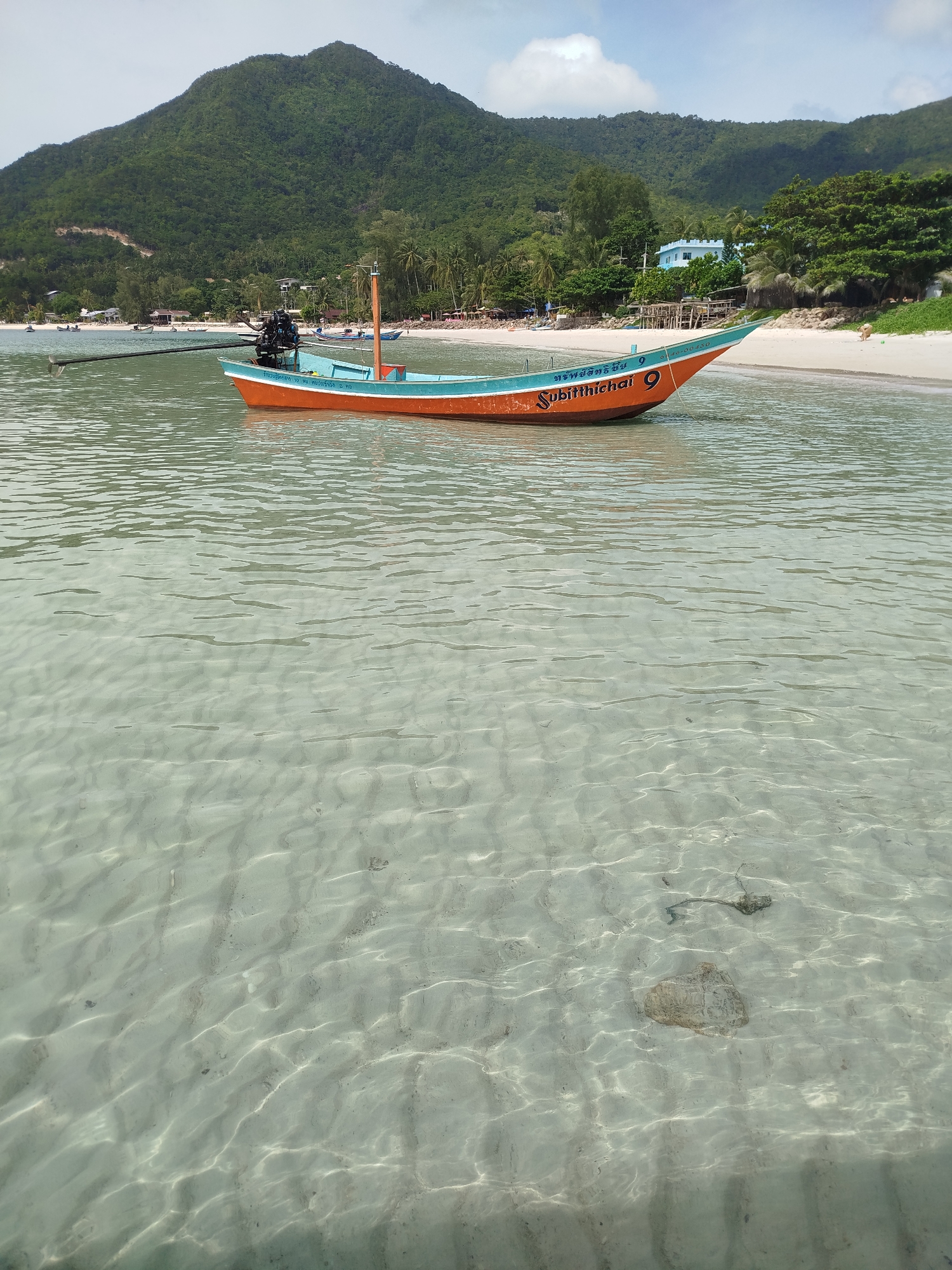 An orange and teal fishing boat with an outboard motor is anchored in shallow, clear water near a sandy beach. The beach is lined with palm trees and lush green hills in the background. Several other boats and a few people are visible on the beach and in the water further down the coast. The water's surface is rippled, and the sandy seabed is visible beneath the clear water. The sky is partly cloudy. This scene is typical of a coastal area in Ko Pha-ngan District, Thailand.