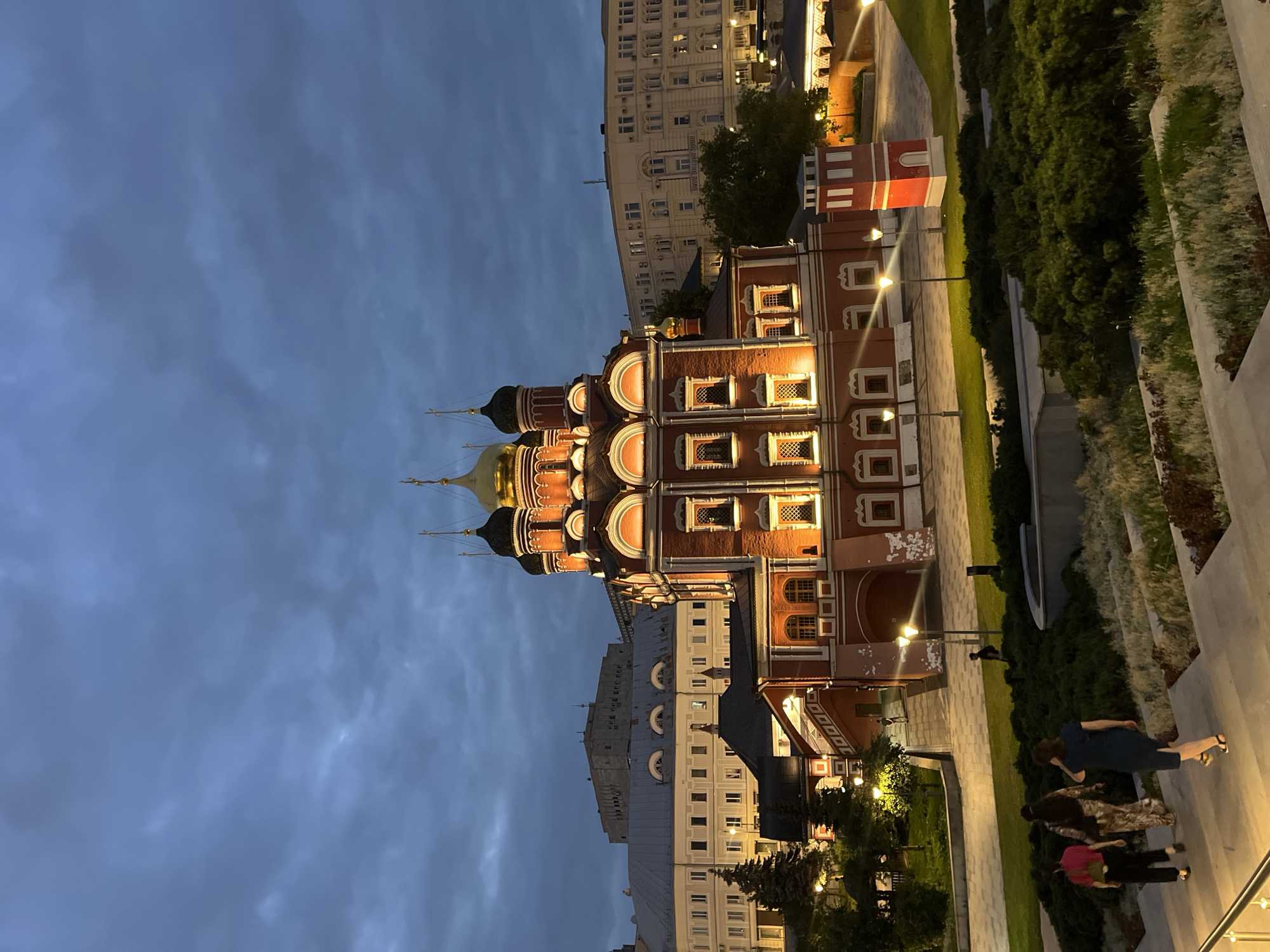 The image captures the Church of the Beheading of Saint John the Baptist in the city of Moscow, Russia, at dusk. The church, constructed of red brick with gold domes, is illuminated by warm interior lights, highlighting its architectural details. In the foreground, three women are walking up a set of concrete stairs, partially obscured by lush green bushes and decorative grasses. Beyond the church, several multi-story buildings, typical of urban Moscow architecture, are visible under a cloudy, dark blue sky. The scene is illuminated by streetlights, casting pools of light on the paved areas and walkways around the church. A small, red, tower-like structure stands to the right of the church.