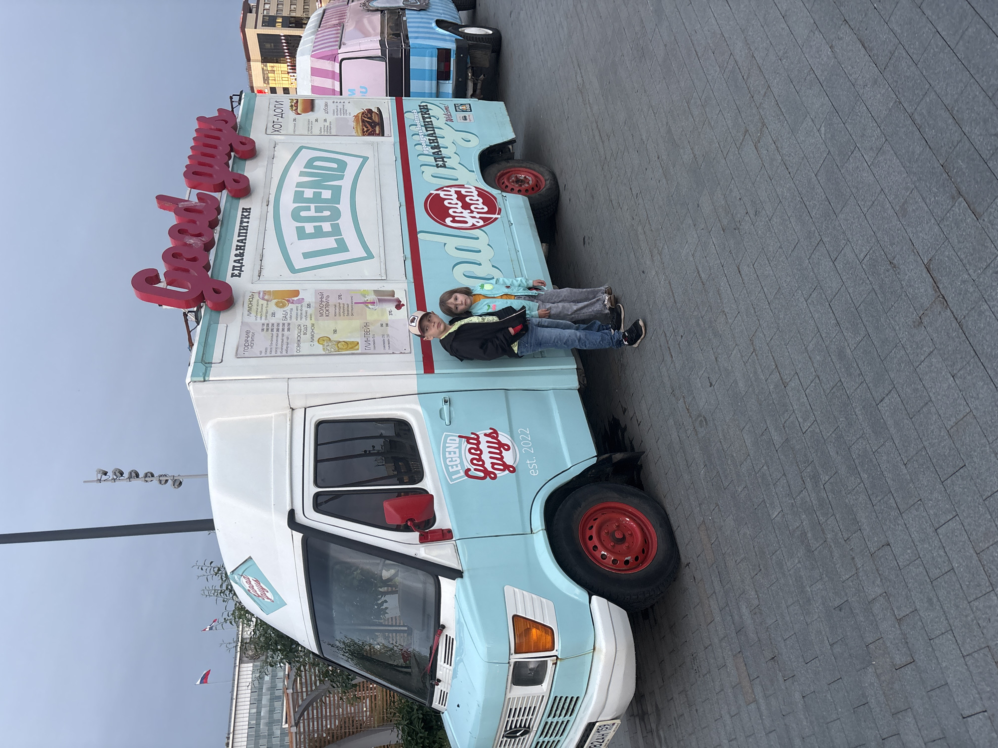The image depicts a paved urban space in Perm, Russia, featuring two food trucks and two children. The foreground is dominated by a light blue and white Mercedes-Benz T2 food truck, identified by its front grille, headlights, and a visible Russian license plate "X824AH 159" which indicates Perm Krai. This truck has "Good Guys" in red script on its roof, "ЕДА&НАПИТКИ" (Food & Drinks) below it, and "LEGEND" in a banner on its side. Menu boards display offerings like hot drinks, lemonades, milkshakes, and hot dogs. The truck's front wheels feature red hubcaps.
Two children stand to the left of the front truck. A boy on the left wears a dark baseball cap, dark jacket, blue jeans, and sneakers, looking towards the viewer. A girl on the right wears a light green jacket, grey pants, and light-colored shoes, also looking towards the viewer.
Behind the first truck, a second food truck is partially visible, featuring pink and white stripes on its upper section and blue and white stripes on its lower section, appearing to be a modified Volkswagen Type 2 (T3) van.
The ground is covered with grey rectangular pavers. The background shows an overcast sky and several multi-story buildings, some with reflective glass facades. A tall, dark street light pole stands prominently, along with another pole equipped with multiple upward-facing floodlights. Two flags are visible above the buildings in the distance: a clear white, blue, and red horizontal tricolor (the flag of Russia), and a second tricolor flag with similar colors but less distinct due to distance and movement. Sparse vegetation, including small trees and bushes, is present behind the food trucks, alongside a wooden slat structure.