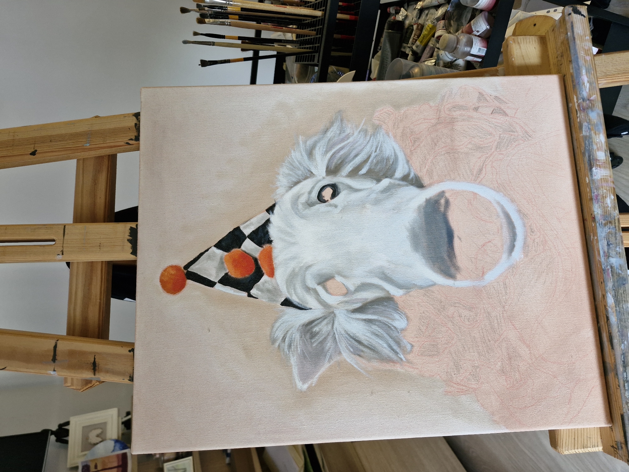 A canvas painting is prominently displayed on a wooden easel, capturing a whimsical scene in what appears to be an artist's studio. The central subject of the painting is a white, furry animal's head, possibly a dog or sheep, depicted wearing a black and white checkered party hat adorned with two bright orange pom-poms. The animal's eye is visible and expressive, and its fur around the face and ears shows detailed texture. A prominent snout or mouth area is also visible.
The painting is clearly in an unfinished state; while the animal's head is largely rendered, the background is a light peachy-pink wash over which faint red pencil sketches outline additional elements or a planned backdrop. The texture of the canvas is visible across the entire surface.
Behind the canvas, numerous artist's paintbrushes are neatly stored in a holder, and various tubes and bottles of paint are visible, reinforcing the setting of an active art workspace. The wooden easel itself bears signs of use, including scattered paint marks. A wooden floor is partially visible, along with a framed picture in the lower left background. The overall impression is one of creative work in progress. No legible text is visible.