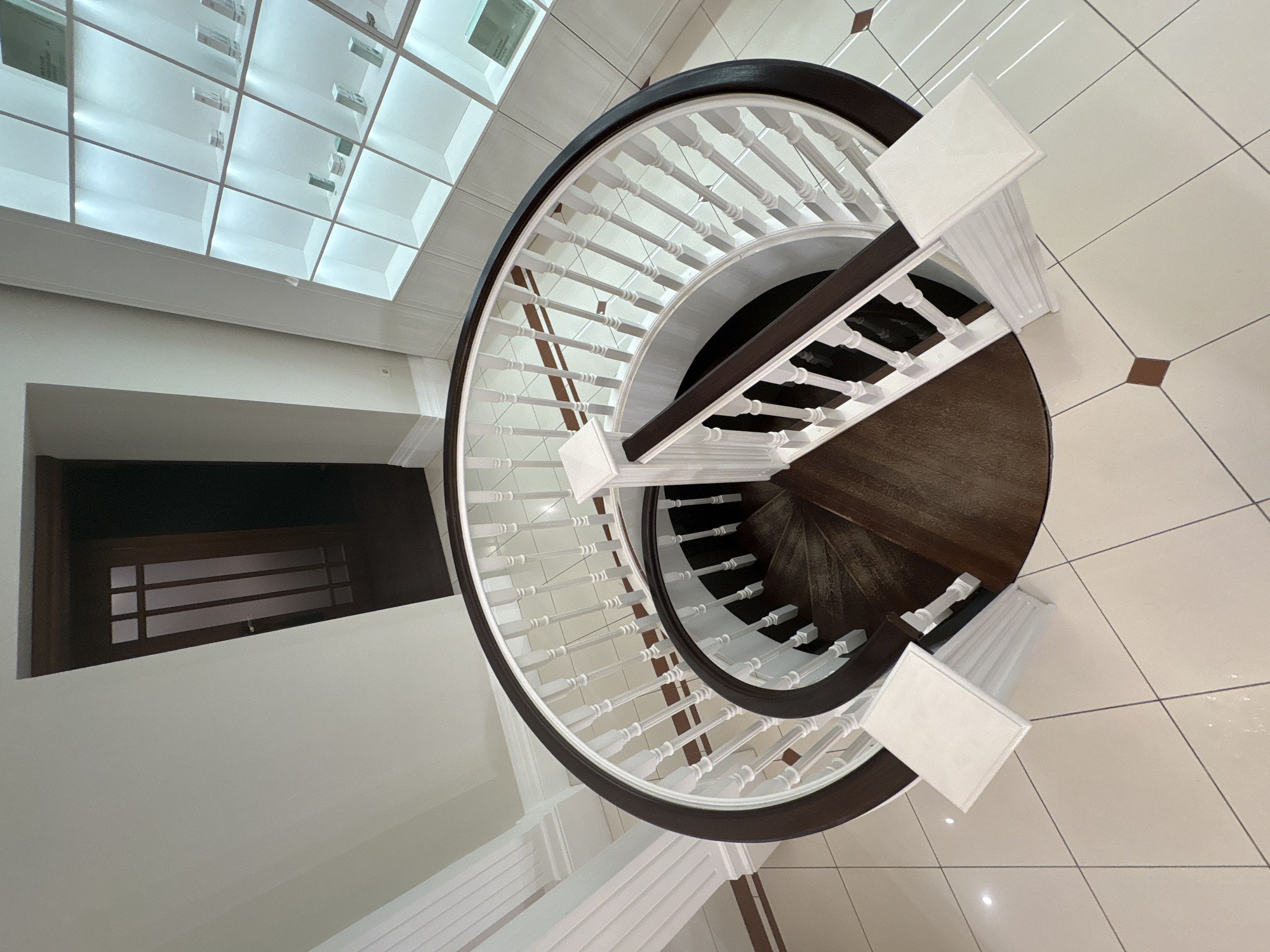 This media file captures a high-angle, top-down view of a spiral staircase within a modern building interior, likely located in Khimki, Russia. The prominent subject is a grand spiral staircase, characterized by its dark brown, elegantly curved banister and numerous light-colored, possibly white, turned balusters and newel posts. The staircase treads are also dark brown, creating a strong contrast with the lighter balustrade.
The surrounding floor is paved with large, light-colored, glossy square tiles, likely cream or off-white, accentuated by dark gray grout lines. A decorative element is introduced through small, dark brown square tiles inlaid periodically into the main flooring.
Above the scene, a striking grid-patterned ceiling feature dominates, appearing to be a large skylight or an integrated lighting system with translucent panels, providing abundant, even illumination. Visible within some of these panels are faint reflections and what appear to be labels or components of building systems; partially legible text includes "MANAGEMENT SYSTEM."
To the left, a dark rectangular opening reveals a dark wooden door with horizontal paneling, suggesting an entrance to an adjacent room or hallway. The walls are painted a uniform light color. There are no people visible, and no specific activity is taking place, presenting a static architectural study. The bright and clean aesthetic, along with the detailed craftsmanship of the staircase, suggests a well-maintained and sophisticated interior.