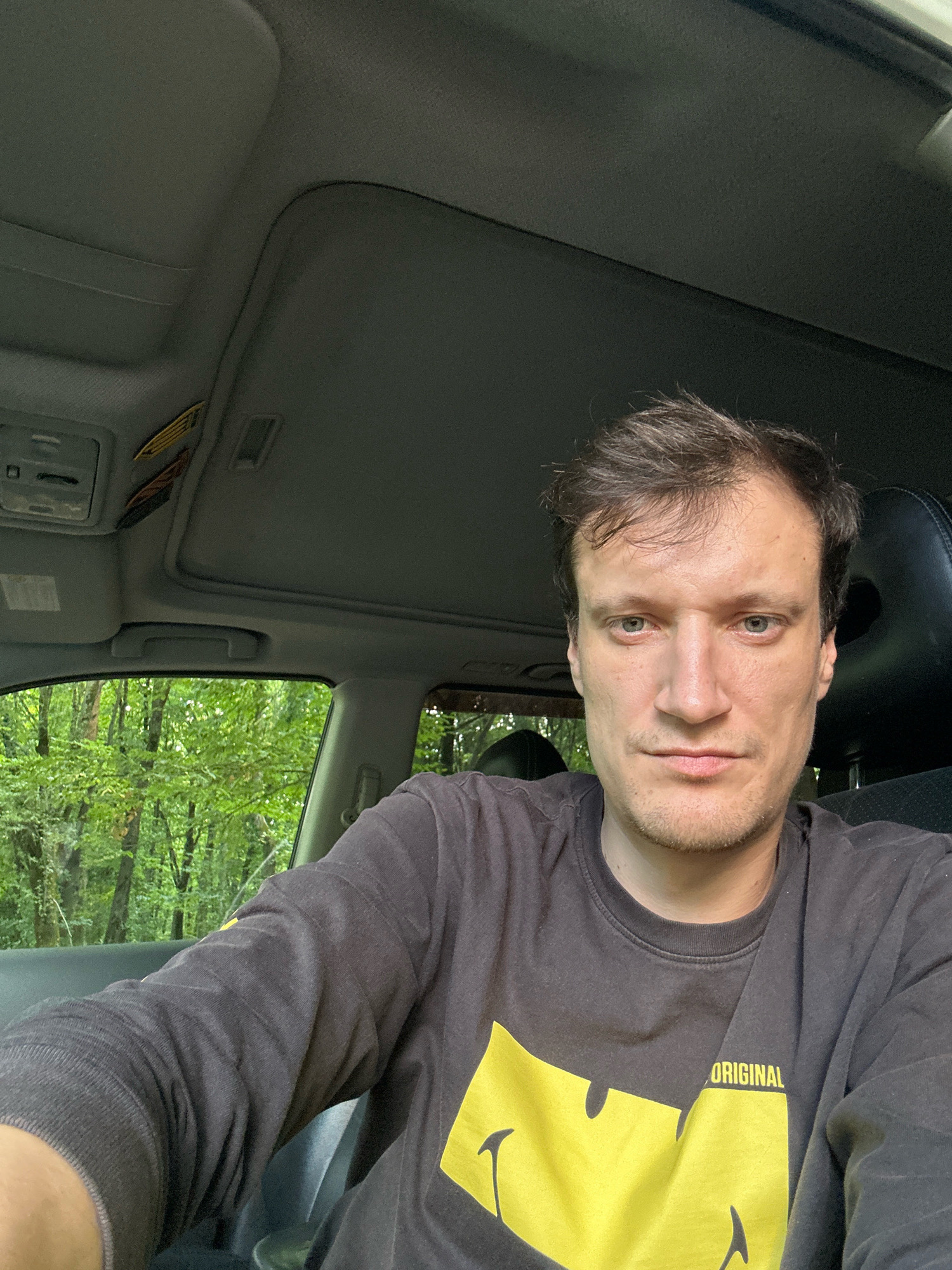 A man with short brown hair and light blue eyes is taking a selfie inside a car. He is wearing a dark gray long-sleeved shirt with a yellow graphic and the word "ORIGINAL" printed on it. The background shows trees and foliage through the car windows, suggesting an outdoor or rural setting. The car's interior, including the roof lining and a portion of the dashboard, is also visible. The image appears to be taken during daylight. The provided crops do not offer any specific information about the location in Shatava, Ukraine.