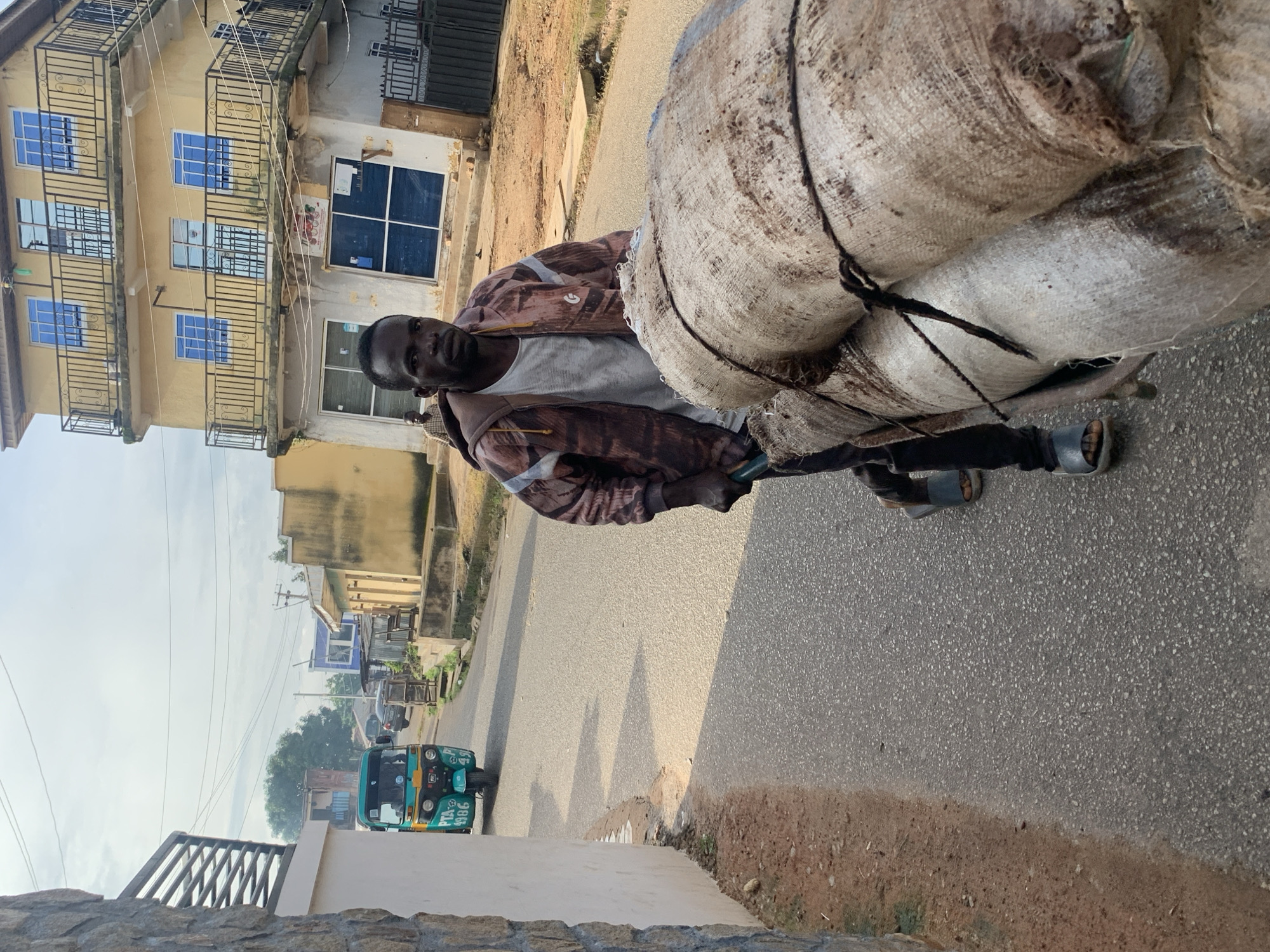 A man in a patterned jacket and dark pants pushes a cart loaded with two large, rough-textured sacks down an asphalt road in Jos, Nigeria. The sacks are tied with dark rope. The man is walking towards the viewer, looking directly at the camera. He is wearing dark sandals. To the left of the man, a light-colored wall and a metal fence are visible. Further down the road, a turquoise and yellow auto-rickshaw with the license plate "PTA 4986" is parked on the left side. In the background, there is a multi-story yellow building with blue windows and balconies, as well as other buildings and trees. The sky is overcast.