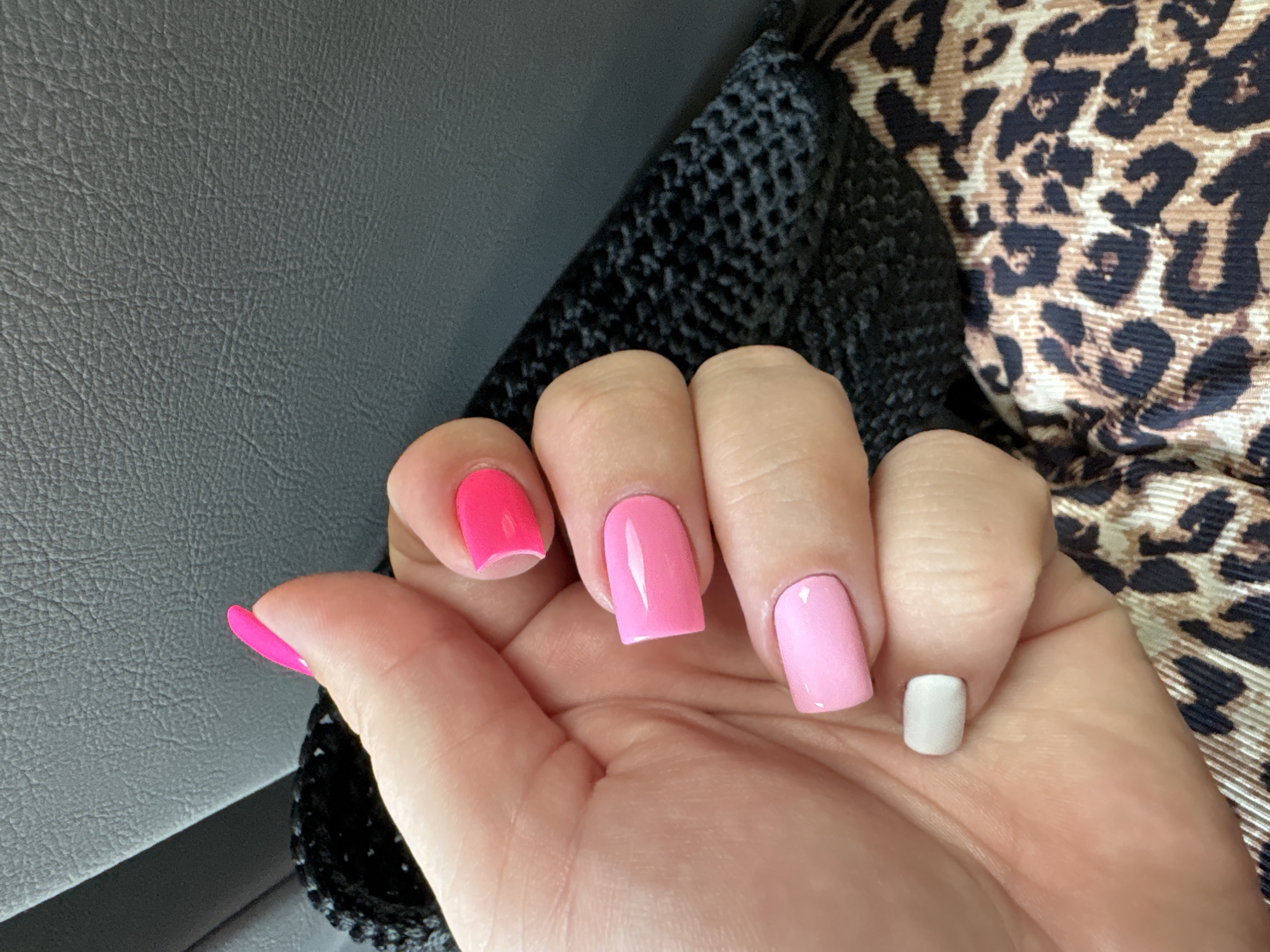 A close-up, overhead shot features a person's left hand with a manicure of different colored nails against a dark gray, textured surface and a black, woven material. The nails are painted in shades of pink, with the thumb nail a bright neon pink, the index finger a hot pink, the middle finger a lighter pink, and the ring finger a pale pink. The little finger is painted a light, muted gray. The hand is positioned as if holding or interacting with the black woven material, which appears to be part of a bag or accessory. Behind the hand and woven material, the dark gray surface has a distinct leather-like texture. The lower right portion of the image shows a piece of fabric with a leopard print pattern in shades of brown and black. The scene appears to be indoors, possibly in a vehicle or seated area, given the textures and lighting. There are no visible landmarks or context to suggest a specific location within Мастоцкі сельскі Савет, Belarus.