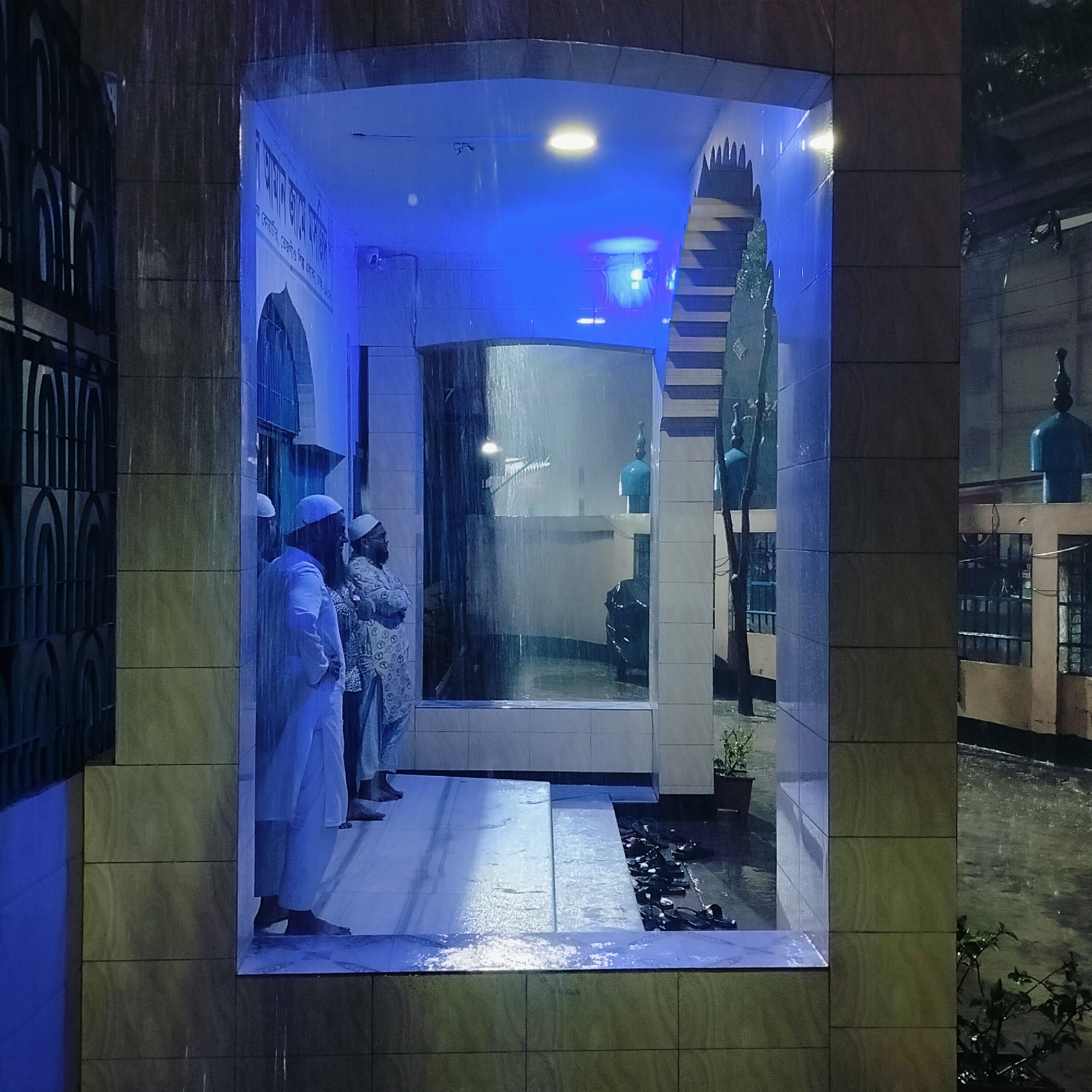 Here's a forensic description of the image:
**Scene:** An outdoor view under heavy rain, likely at night given the prominent artificial lighting. The primary subject is an open structure with tiled walls, resembling an entrance or a window-like opening.
**People:** Two men are standing inside the structure. Both are wearing what appears to be traditional Islamic attire, including head coverings. They seem to be observing something outside.
**Objects:** Within the structure, there is a water feature resembling a thin curtain of rain or a fountain. Outside the structure, there are buildings, plants in pots, and what appears to be a rack of shoes. The presence of domes further suggests that the location may be affiliated with a mosque or Islamic institution.
**Actions:** The men are standing and observing. The rain is falling heavily.
**Interactions:** The people are interacting with each other.
**Environment:** The scene is dominated by the rain and the artificial blue lighting of the room. The general atmosphere is wet and possibly cold. Given the presence of traditional Islamic attire and architecture, it is plausible that this is in Dhaka, Bangladesh.