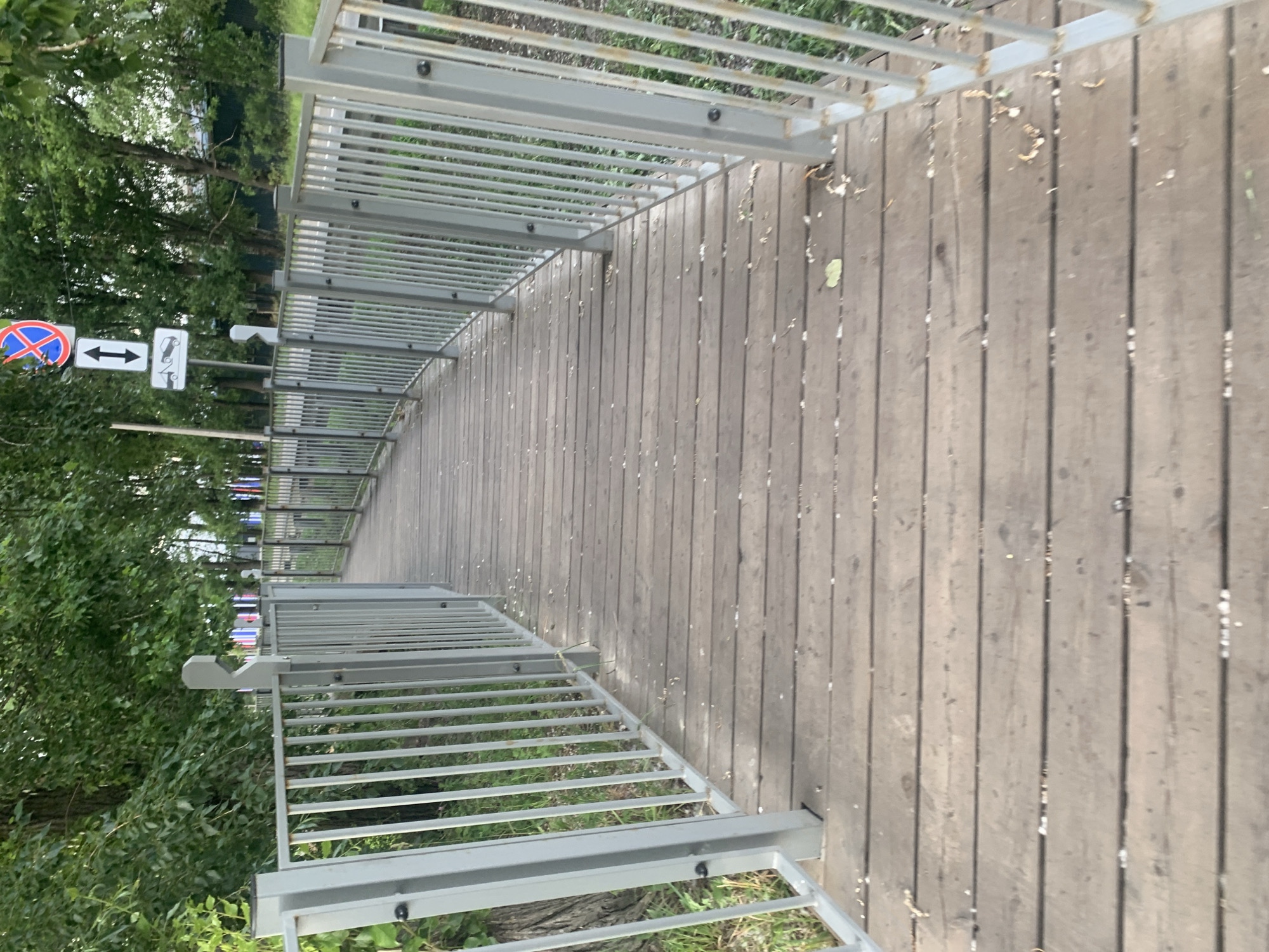 The image shows a wooden pedestrian bridge with metal railings. The bridge is situated alongside a tree-lined area and appears to be in a park or natural setting. The perspective is from the bridge, looking down its length. The wooden planks of the bridge show signs of wear and weathering. The metal railings are a light gray color and have a simple, functional design.
To the left of the bridge, there are several traffic signs. One is a circular red and blue sign with a diagonal line across it, indicating a prohibition. Another sign is rectangular and white with black arrows pointing in opposite directions, suggesting a two-way path or movement. A third sign, also white with black graphics, depicts a silhouette of a car with a line through it, possibly indicating no parking or no vehicles. The presence of these signs suggests the bridge is located near a road or a public thoroughfare.
The surrounding greenery indicates it is daytime, likely with overcast or diffused lighting, as there are no strong shadows. The leaves on the trees are full and green, suggesting a warmer season like late spring or summer. The overall atmosphere of the scene is calm and ordinary, depicting a typical outdoor public space. There is no discernible activity or people visible in the image. The location context provided is Kolpino, Russia.