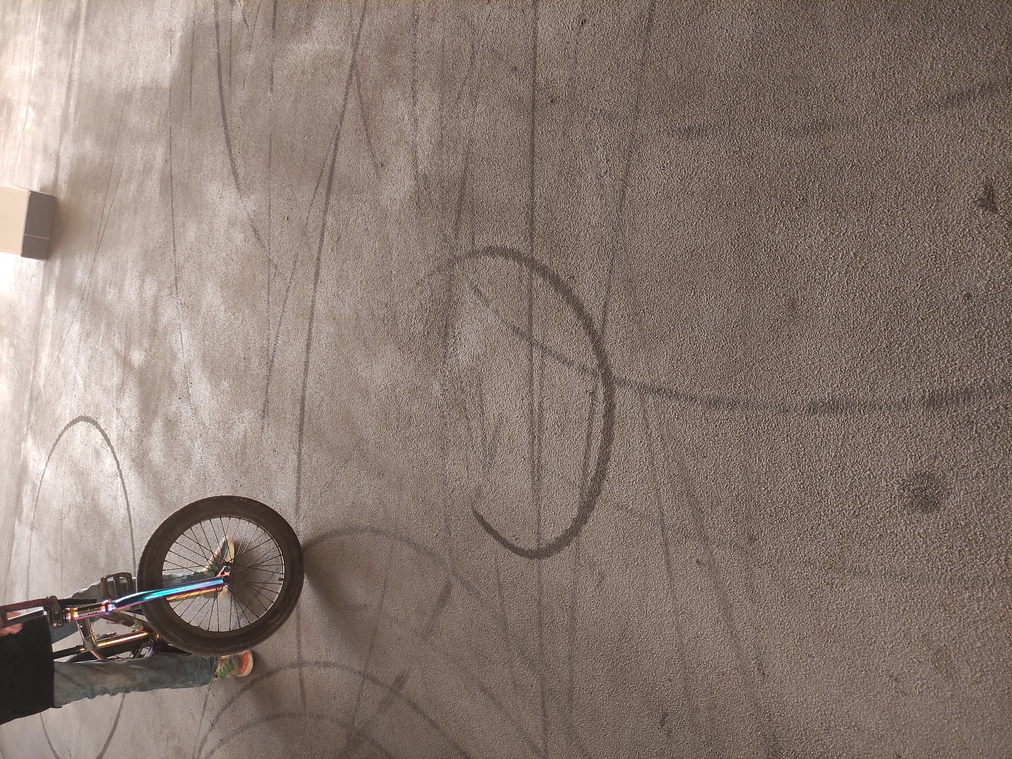 A high-angle shot shows a bicycle on a concrete surface with circular tire marks. The bicycle's front wheel is visible in the lower left corner, with an iridescent frame and multicolored spokes. A person's leg, wearing jeans and a multicolored shoe, is partially visible near the bicycle. The tire marks consist of several overlapping circles, some of which are more distinct than others. A rectangular object is present in the upper left corner. The scene is illuminated, but the source of the light is not visible. The location appears to be an urban or industrial setting, characterized by the bare concrete surface.