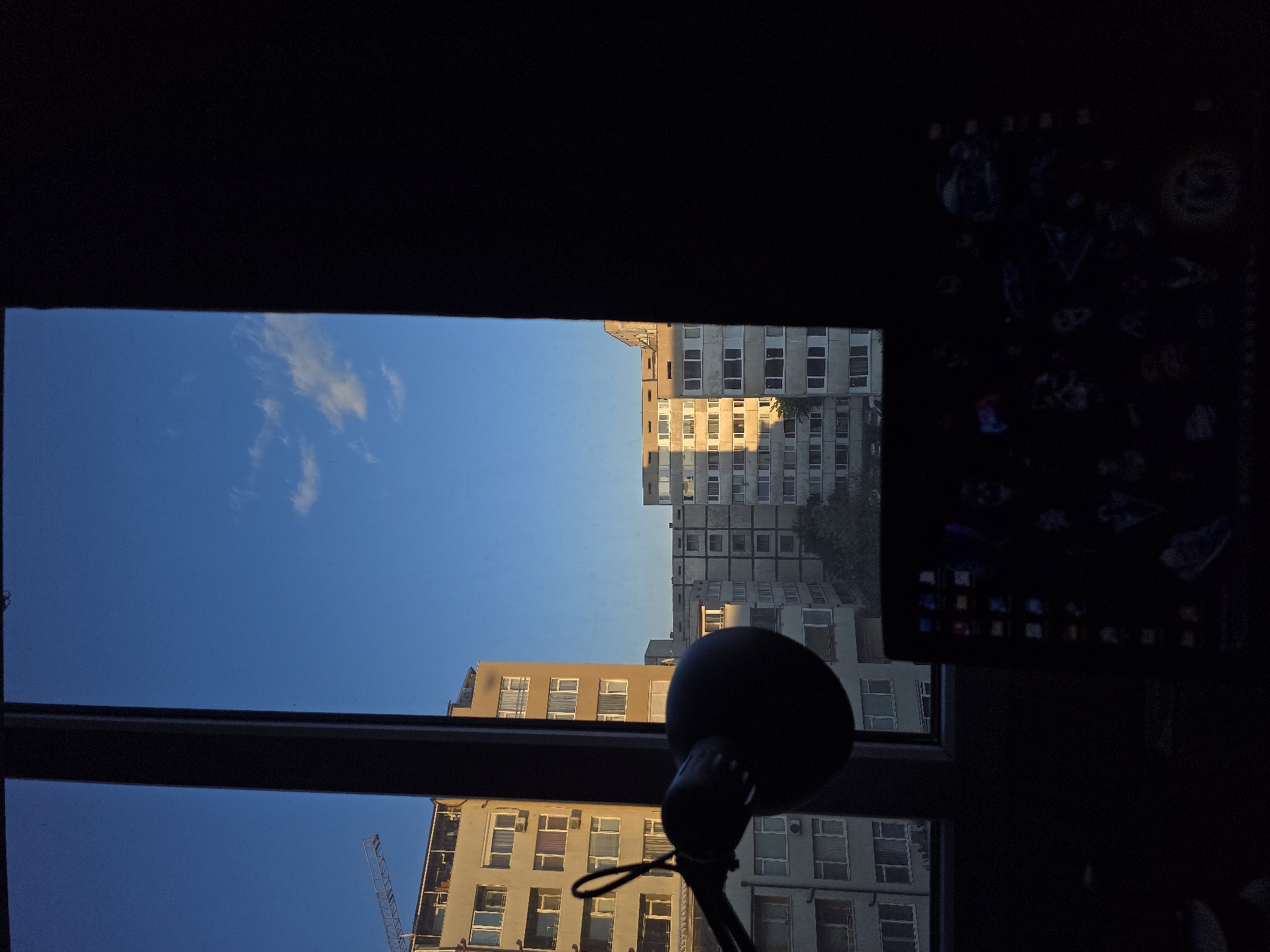 The image depicts a view from a window in Sofia, Bulgaria. Outside the window, several apartment buildings are visible, illuminated by the setting sun. The sky is blue with scattered clouds. A construction crane is partially visible in the lower left corner. An object resembling a lamp is placed in the foreground. A dark computer monitor occupies the right side of the frame. The time of day appears to be late afternoon or early evening.