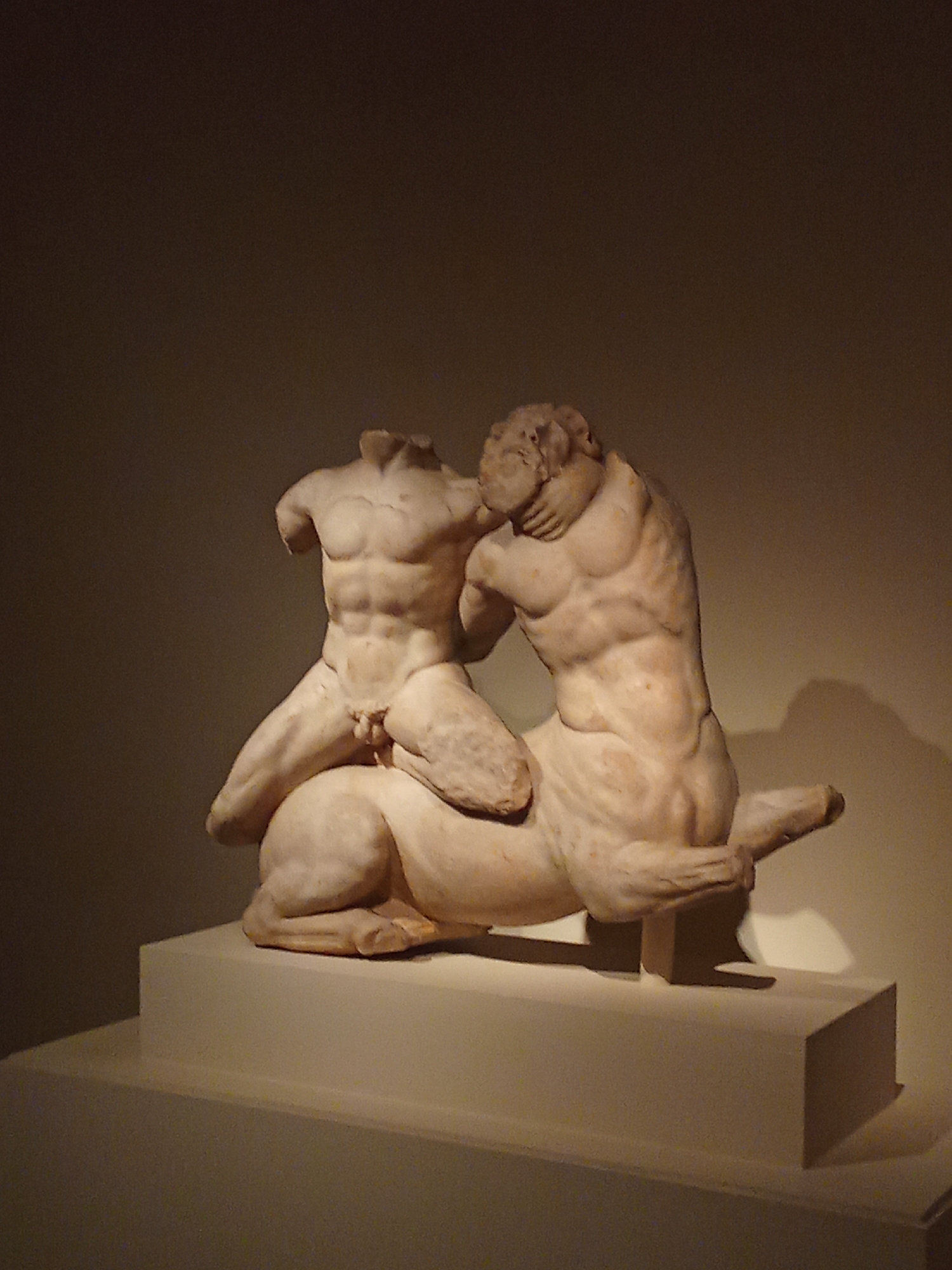 The image shows a white marble sculpture of two figures, likely a centaur and a human, in a dynamic pose, placed on a white pedestal. The figures are headless and appear to be engaged in a struggle or embrace. The sculpture is illuminated by a spotlight from above, creating strong shadows on the background wall and the pedestal. The background is a plain, dimly lit wall. The style of the sculpture suggests classical antiquity. This artwork is located in the Museo Nacional de Bellas Artes in Buenos Aires, Argentina.