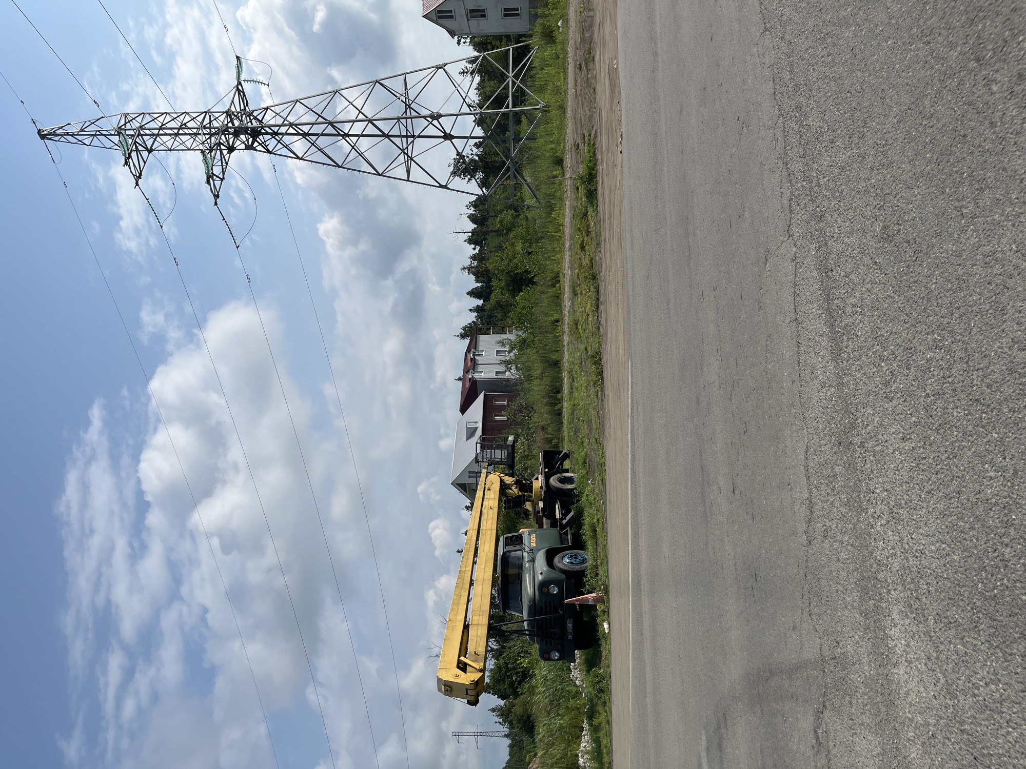 In Ureki, Georgia, a weathered, dark green truck equipped with a long, bright yellow boom crane is parked on the side of a paved road. The truck has blue rims on its rear wheels and a license plate that reads "AB 977-IB". An orange traffic cone sits near the front of the truck. Behind the truck, a dense line of green trees separates the road from several buildings, including a white and red structure and a taller, white building with a red roof. To the right of the buildings, a massive metal electricity pylon dominates the sky, with power lines stretching horizontally across the frame. The sky above is a bright blue, dotted with scattered white clouds. The ground in front of the truck is a mix of overgrown grass and bare dirt, leading up to the road's asphalt surface, which shows signs of wear and tear.