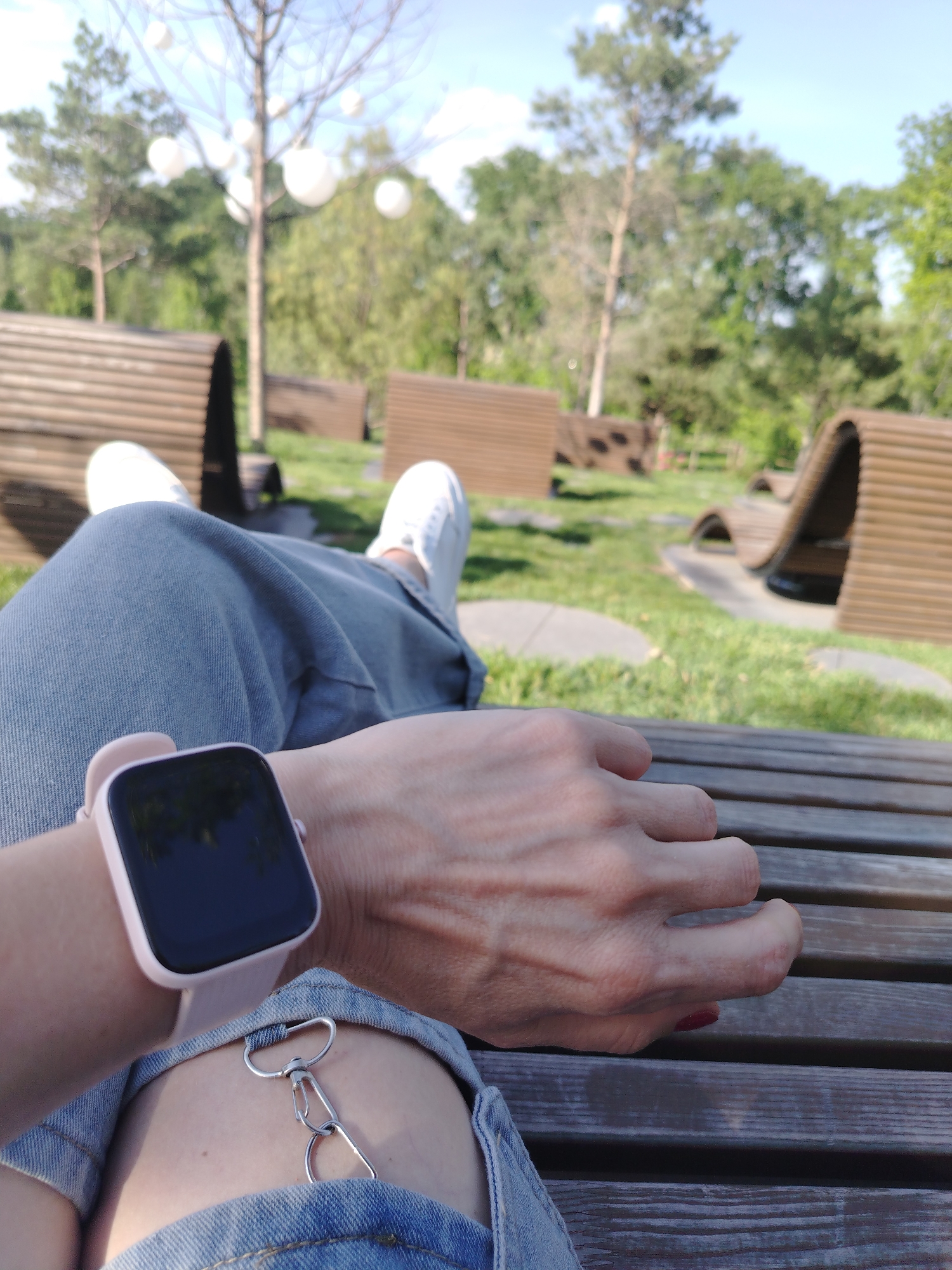 A first-person perspective shot captures a person's legs and wrist resting on a wooden bench in a park. The person is wearing light blue denim jeans, white sneakers, and a pale pink smartwatch. A silver chain accessory is attached to the jeans. In the background, a grassy area is dotted with modern, curved wooden seating structures and trees. The scene suggests a relaxed outdoor setting, possibly in a public park within the city of Krasnodar, Russia, given the context provided.