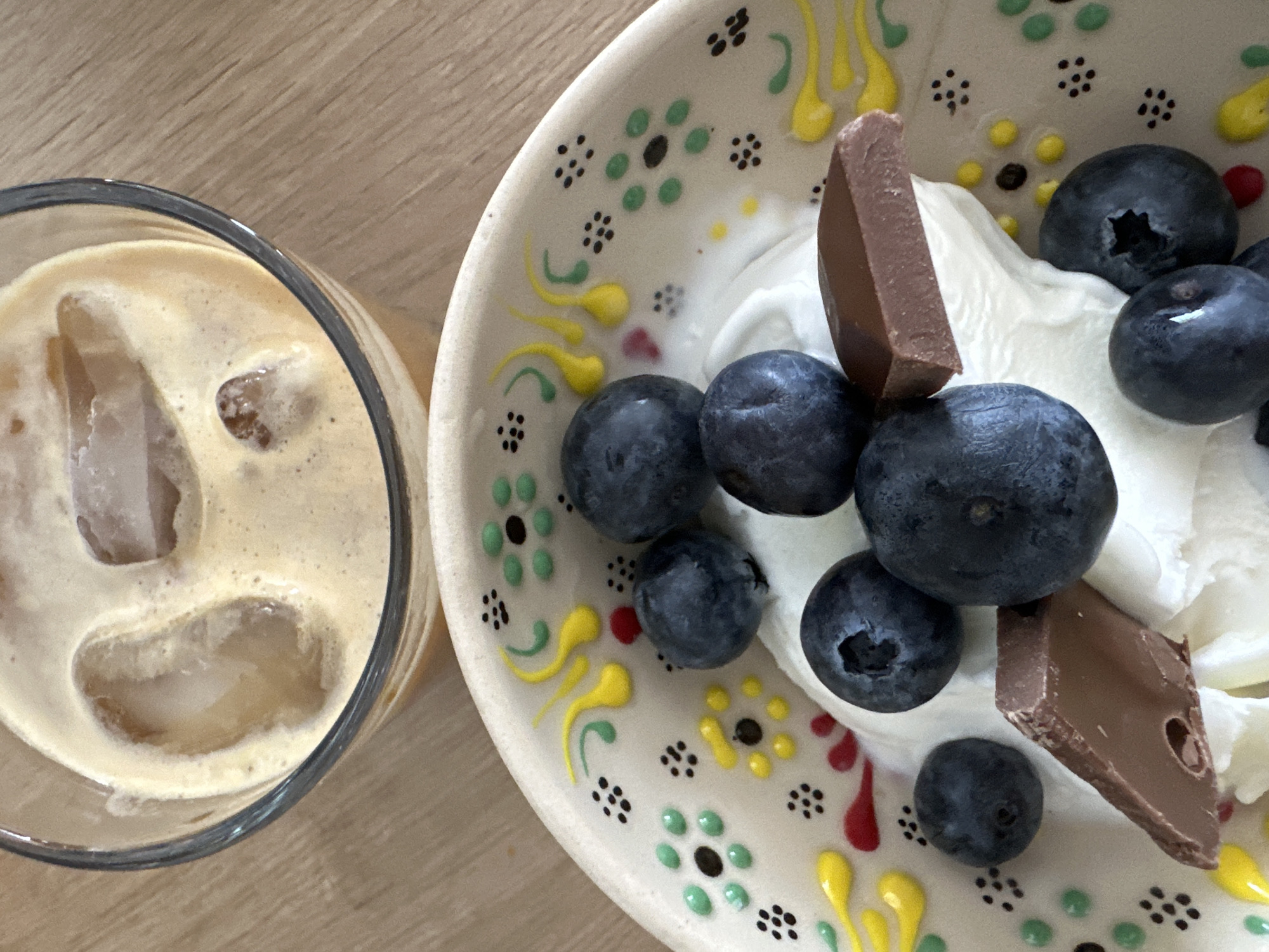 The image presents a top-down view of an iced beverage and a dessert bowl, both positioned on a light-colored wooden surface. The upper section of the frame shows a transparent glass containing a light brown, frothy iced drink with visible ice cubes. In the lower section, an off-white ceramic bowl features a hand-painted rim adorned with green dot-flowers, black dot clusters, yellow swirls, and red accents. The bowl's contents consist of a white, creamy substance topped with numerous dark blue blueberries and several broken pieces of milk chocolate. This scene is located in Fethiye, Turkey.