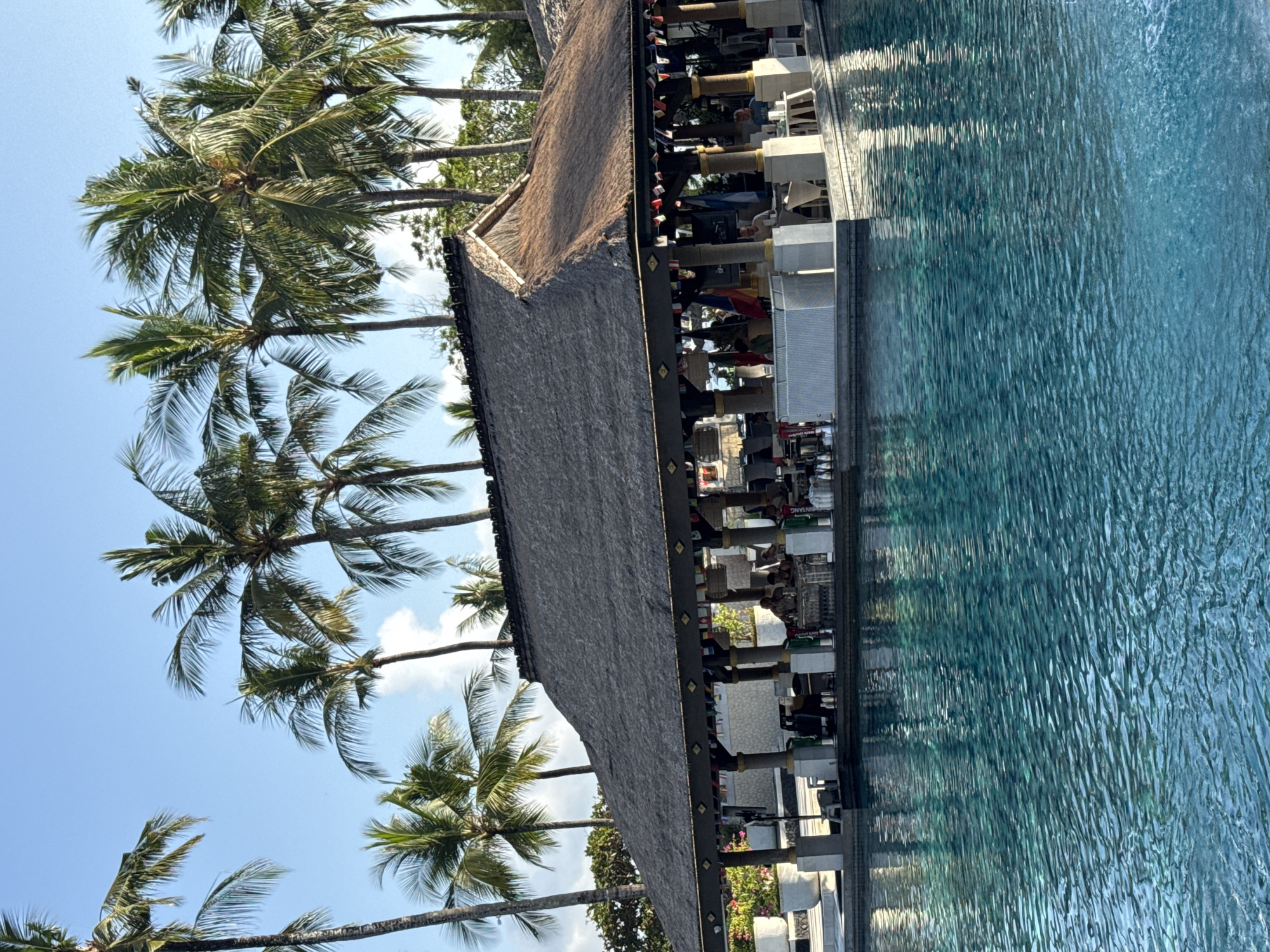 This image depicts a tropical resort bar situated by a swimming pool in Kuta Selatan, Indonesia. The bar features a thatched roof and is decorated with small flags from various countries. Several patrons are seated at the bar and at tables within the structure. Tall palm trees surround the area, with a clear blue sky visible above. The water in the pool is a vibrant turquoise, reflecting the surrounding environment and the clear sky.