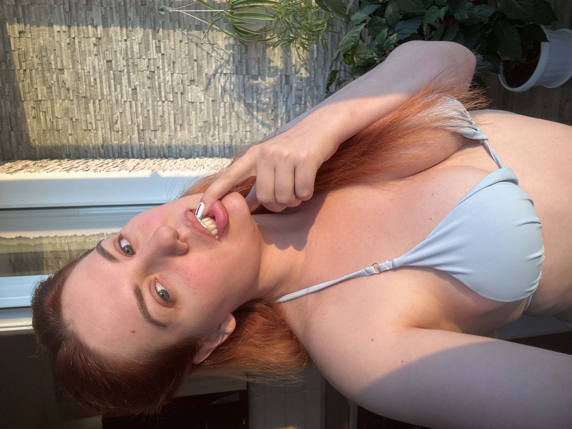 A medium shot shows a woman with reddish-brown hair and fair skin wearing a light blue bikini top. Her left hand is raised, with her index finger touching her lips, and she appears to be biting down on it. She is looking towards the camera with a slight smile, and her eyes are open. Her teeth appear to have some sort of white attachment. The background consists of a stone-tiled wall and a green plant in a pot. The lighting suggests it is either sunrise or sunset, casting long shadows and warm tones on the scene. The location is unknown.