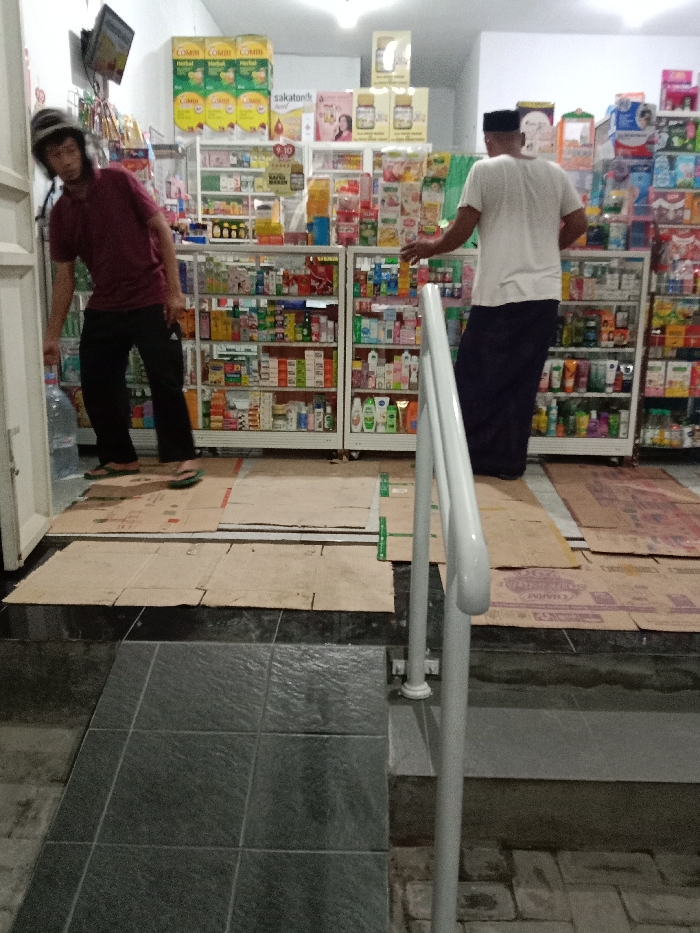 The image displays the interior of a retail establishment, identified as being in the city of Turunsih, Indonesia, observed from a low angle near the entrance. Two adult males are present inside the store. On the left, a male wearing a dark helmet, maroon short-sleeved t-shirt, and dark athletic pants, with green flip-flops on the floor beside his bare feet, leans forward, handling a large clear plastic water bottle positioned near the doorway. On the right, a second male, attired in a white short-sleeved t-shirt and dark sarong-style trousers or pants with a dark cap, stands with his back mostly to the camera, reaching towards a white metal display shelf.

The store's interior features several white metal display shelves densely stocked with various packaged consumer goods, including health supplements (e.g., "COMBI Herbal," "sakatonik"), personal care products, and other merchandise. The floor within the store is largely covered with flattened cardboard boxes, secured in places by small green clips. The entrance area transitions to dark gray tiled flooring, which includes a textured ramp section in the lower left foreground. A prominent white metal handrail is situated in the foreground, indicating access into the store. A small television screen is mounted high on the wall to the left. The scene is illuminated by overhead fluorescent lighting.
