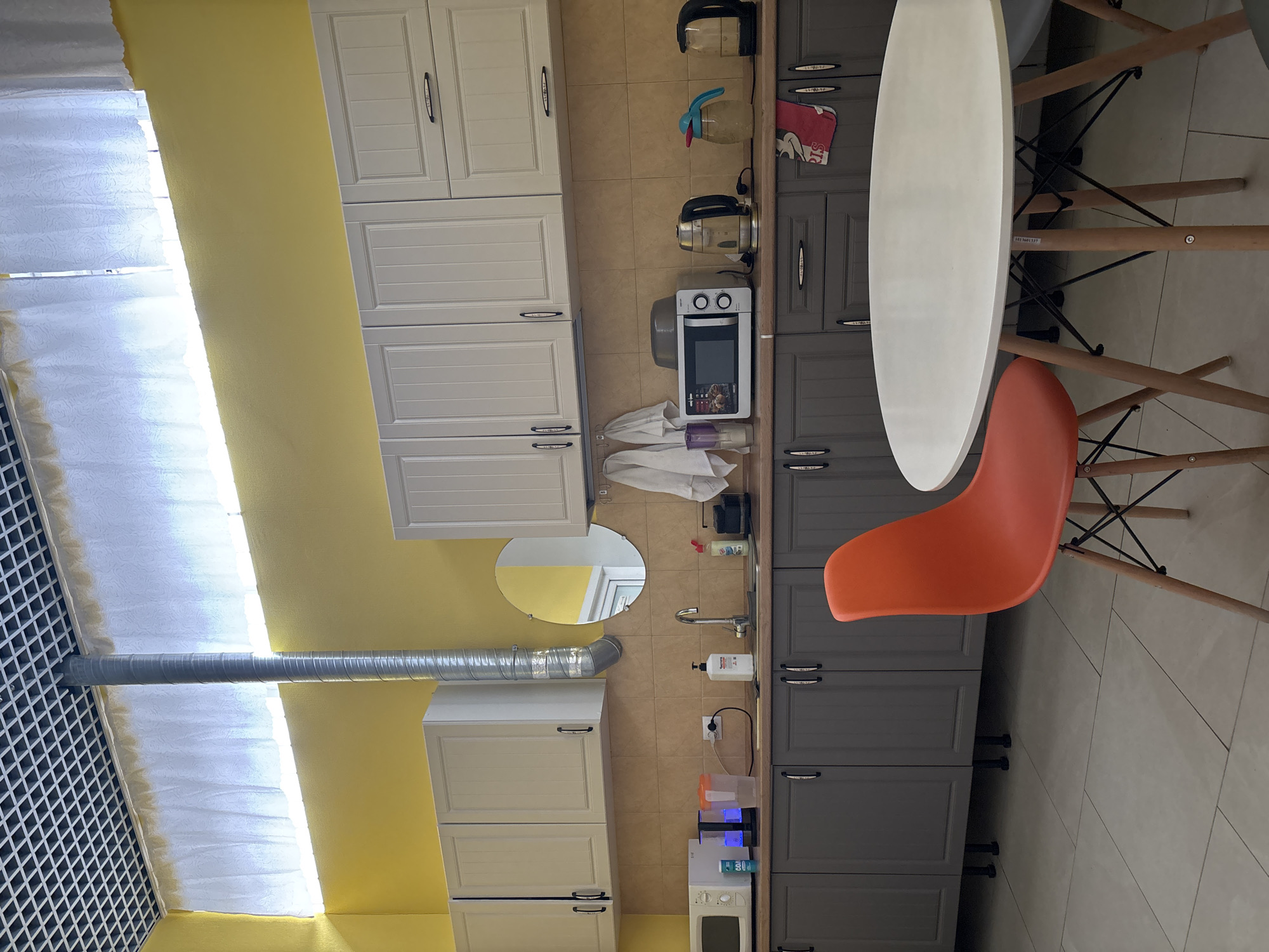 The image shows a kitchen. On the left side of the image, there is a wall painted yellow, and in front of the wall, there are white cabinets. A grey ventilation pipe runs horizontally across the wall. In the center of the image is a sink and countertop. There are grey cabinets below the counter and white cabinets above. On the right, a white table with wooden legs and an orange chair is visible. The location appears to be Saint Petersburg, Russia, based on the style of the kitchen and the building design. The scene contains no people, focusing on the static arrangement of the kitchen interior.