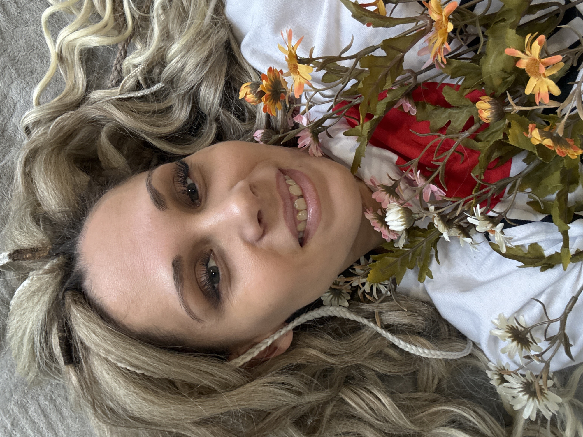 A woman with blonde, wavy hair and braided accents lies on a gray surface. She wears heavy mascara and is smiling, revealing her teeth. She is adorned with flowers, including orange, pink, and white varieties, along with green leaves. She is wearing a white garment, with a hint of red visible. The location is unspecified.