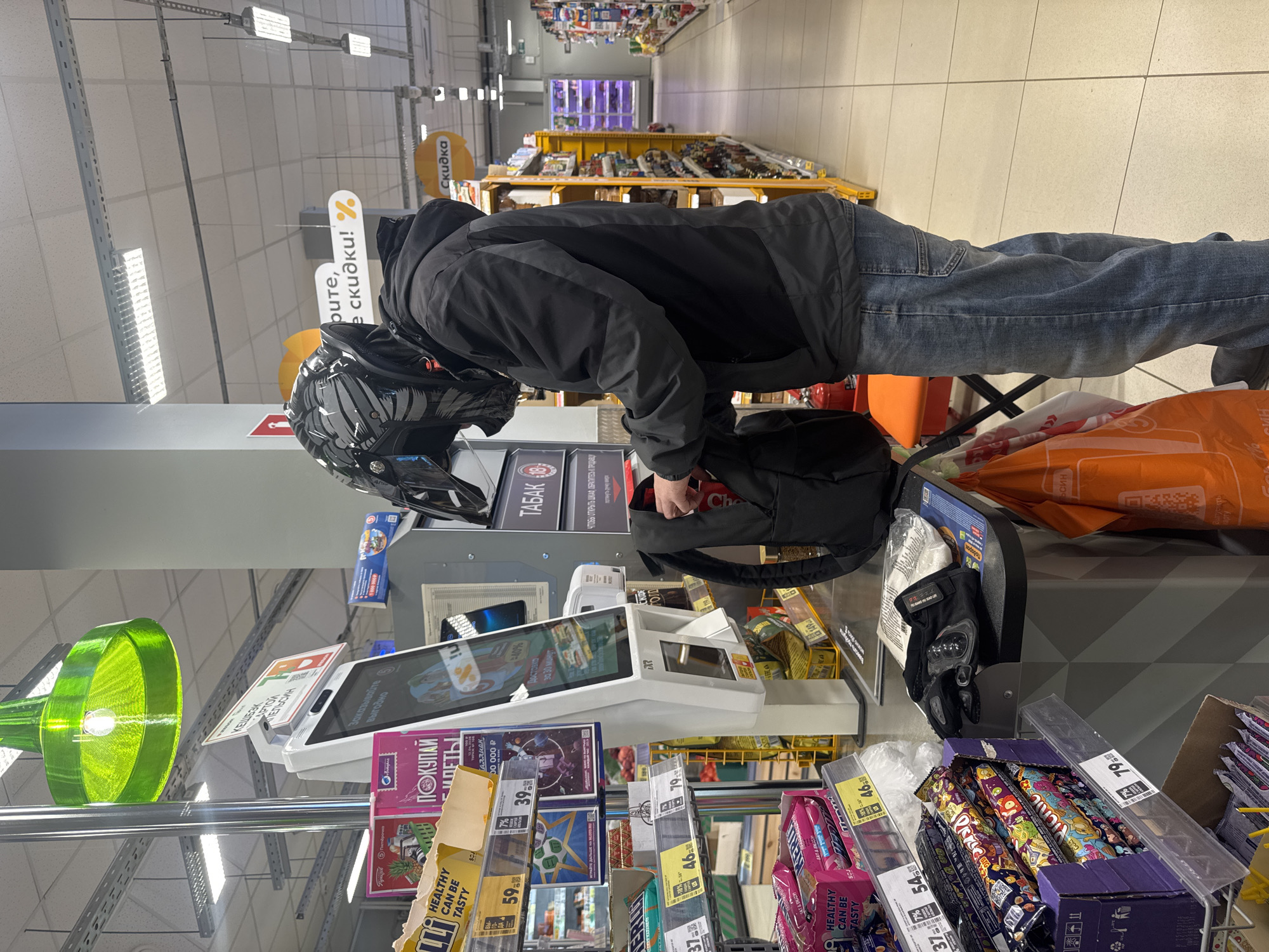 The image captures a person wearing a motorcycle helmet and black jacket bending over a self-checkout station inside a supermarket. The individual is placing items from a black backpack onto the station's counter. The checkout station is surrounded by various items for sale, including candy bars and cigarettes. The environment appears to be well-lit with fluorescent lighting, and the supermarket layout features shelves stocked with goods in the background. The location is identifiable as a "Pyaterochka" supermarket in the city of Chaikovsky, Russia.