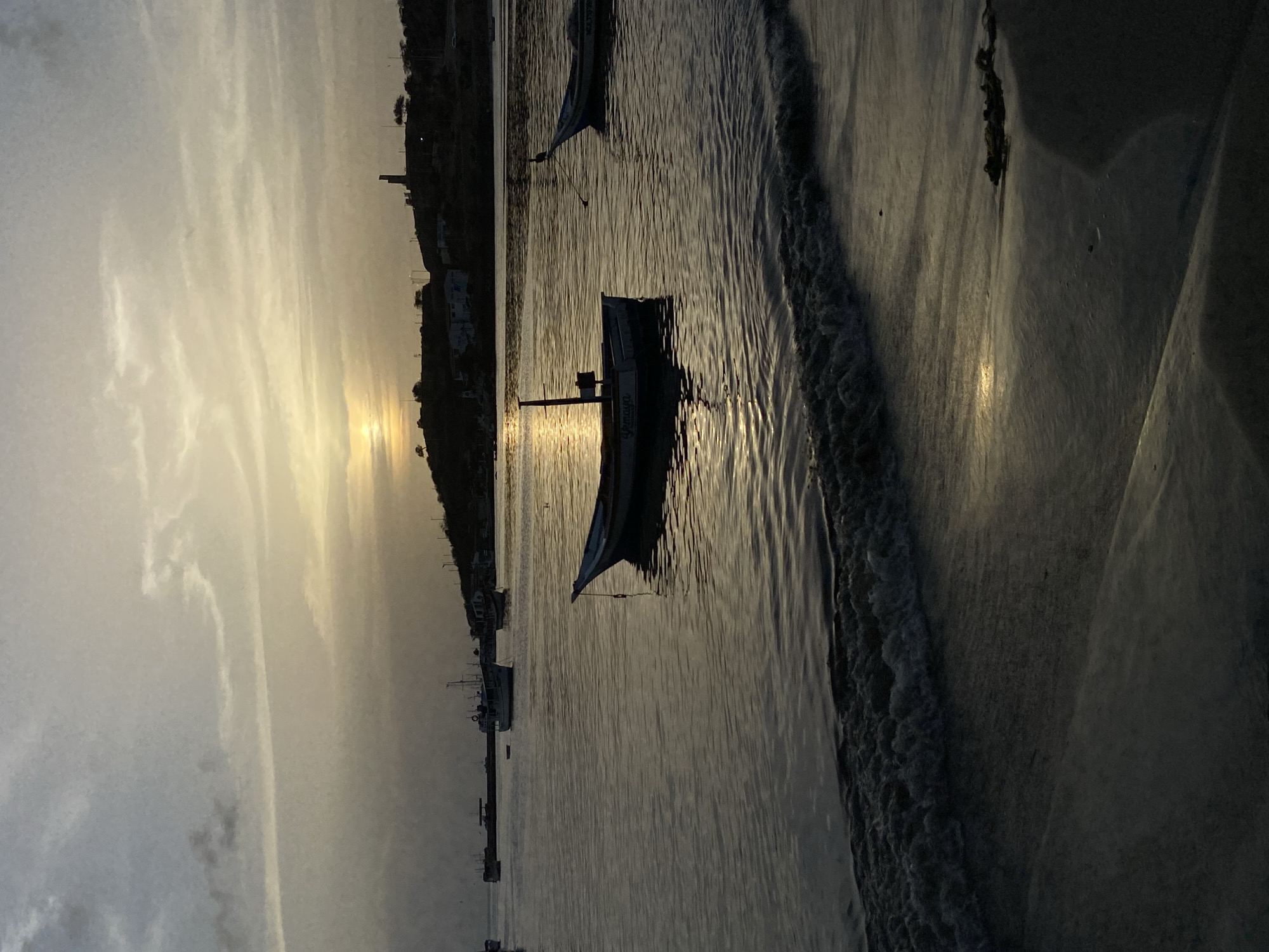 The image depicts a tranquil sunset scene at the coast of Juan Griego, Venezuela. The sky is filled with muted, hazy clouds, with the sun peeking through, casting a warm, golden glow across the water and the horizon. In the foreground, a gentle wave laps onto the dark sand of the beach. The calm sea is dotted with several small, silhouetted fishing boats, some of which appear to be at anchor. In the background, a hilly coastline is visible, with a few structures and a distinct tower-like monument silhouetted against the fading light. The overall atmosphere is serene and peaceful, capturing the quiet beauty of a coastal sunset.