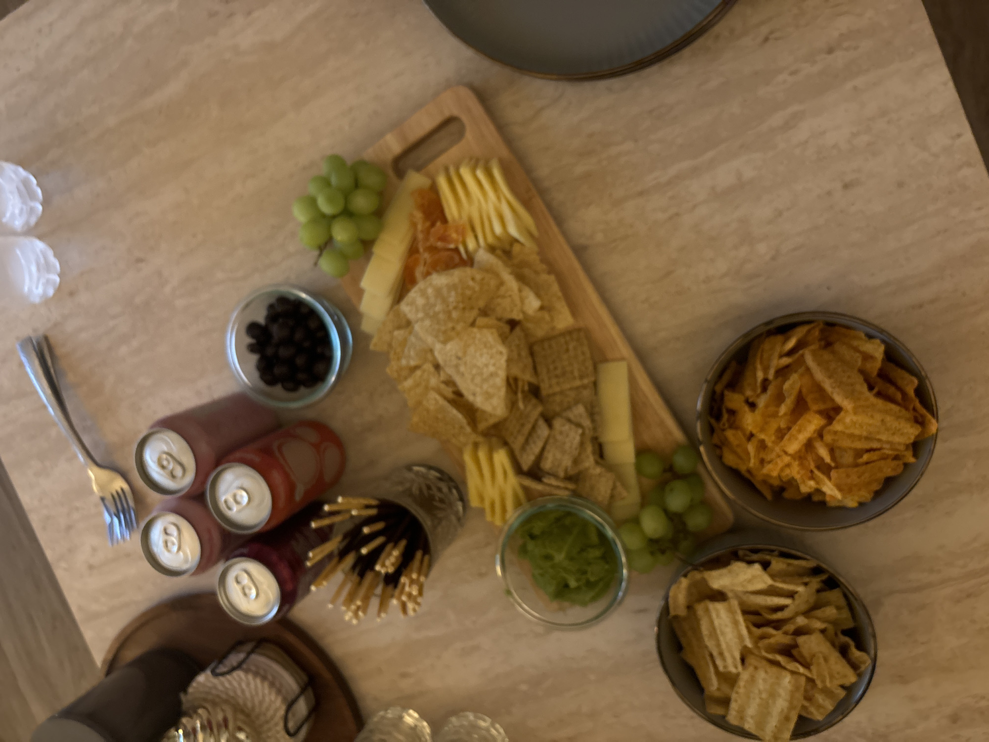 This is a top-down, medium shot of a food spread on a marble-patterned table, likely in a home environment in Hallandale Beach, Florida, United States of America. The arrangement includes a wooden cutting board laden with various snacks. On the board, there are several types of crackers, including rectangular and square varieties, alongside slices of cheese and what appear to be dried fruit or jerky strips. In front of the cutting board, a small glass bowl contains a green dip, possibly guacamole, and a bunch of green grapes. To the left of the board, a tall glass contains a bundle of slender, stick-like snacks, some with dark chocolate coating. Beyond these, several cans of soda are arranged, appearing to be red, pink, and light red. A glass bowl filled with dark, small spherical objects, likely olives or dark berries, is situated near the cans. A fork rests at the top of the image, pointing towards the cans. Two clear plastic cups are also visible near the top right corner. In the lower portion of the frame, two separate bowls are filled with different types of chips; one contains flat, rectangular chips, and the other is filled with ridged, orange-colored chips. A dark gray plate is visible on the right edge of the image. No people are present in the scene, and no specific actions are depicted beyond the static arrangement of food items. The lighting is moderate, casting some shadows and highlighting the textures of the food and table.