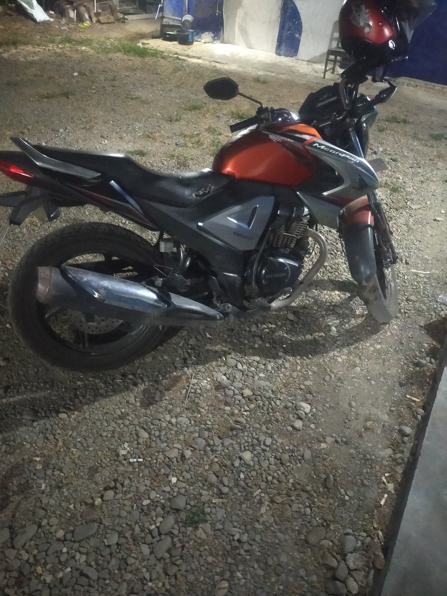 A parked orange and black motorcycle is visible outdoors in what appears to be a dimly lit area with gravel and some dry grass. The motorcycle is positioned at an angle, with its left side facing the camera. A red helmet with a floral design is resting on the handlebars. The background shows some indistinct structures and a blue and white wall. The scene is likely located in Pagerbarang, Indonesia.