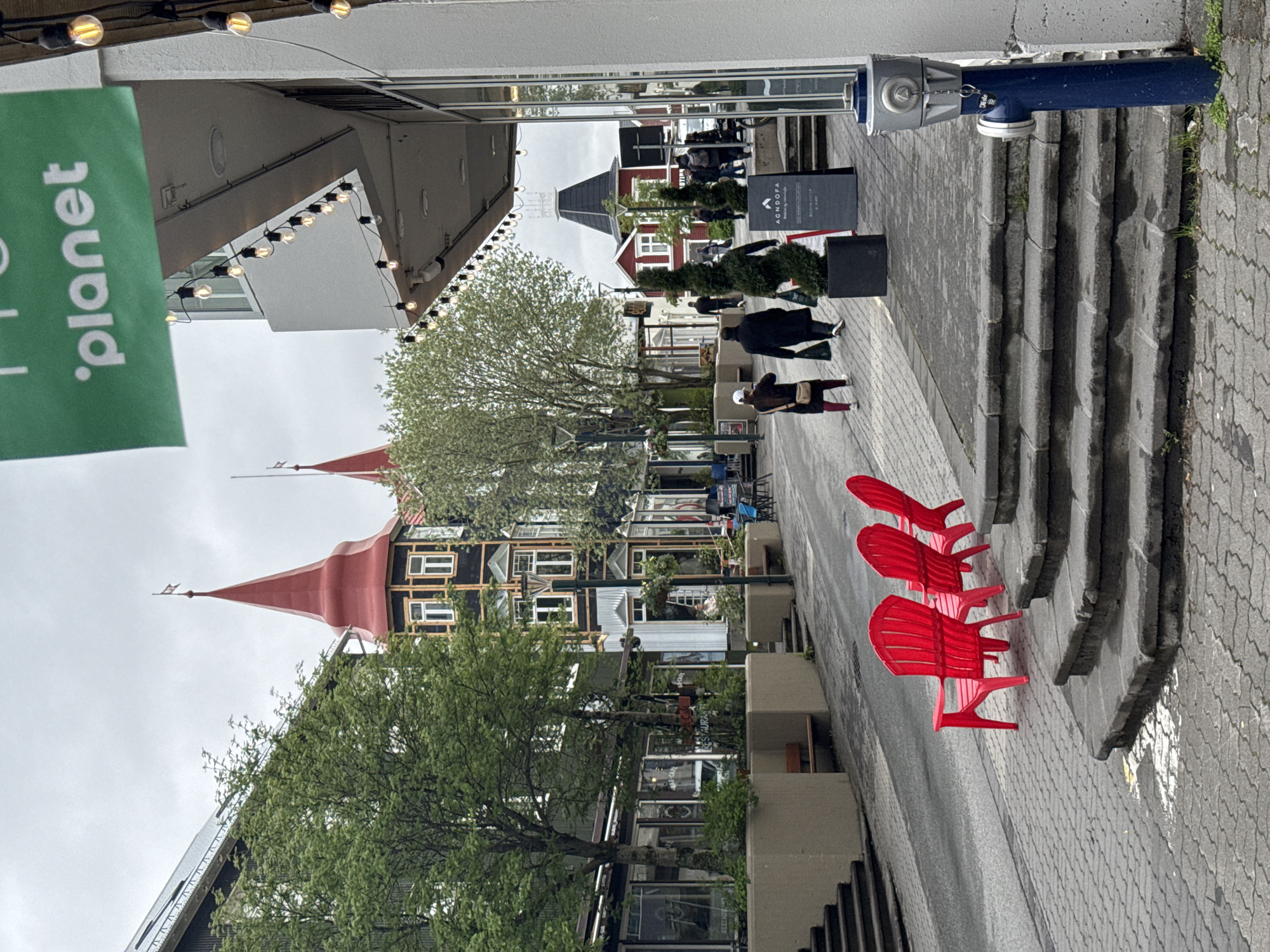 This image captures a street scene in Akureyri, Iceland, under an overcast sky. The view is looking down a pedestrian-friendly street lined with buildings of varying architectural styles.
In the foreground, three bright red Adirondack-style chairs are prominently positioned on the paved sidewalk, facing away from the viewer. To the right, a set of concrete steps leads upwards, and a blue utility pole, possibly a fire hydrant, is visible next to the stairs.
Several people are seen walking along the street further back, appearing as casual pedestrians. The buildings flanking the street feature distinct characteristics; some have dark facades with white window frames, while others boast striking red pointed roofs or steeples, giving the area a unique European town aesthetic. Green trees with fresh foliage are interspersed among the buildings, adding natural elements to the urban setting.
Overhead, strings of warm-toned bulb lights are strung across the street, suggesting an area that might be illuminated in the evenings or designed for a festive atmosphere. A large green banner hangs to the left, partially visible, displaying the text ". planet". Another black sandwich board sign further down the street clearly shows "AGNOOFA" in white lettering, with some smaller, illegible text below. On the blue utility pole, the word "DELTA" is partially visible. The overall impression is a charming and lively town street.