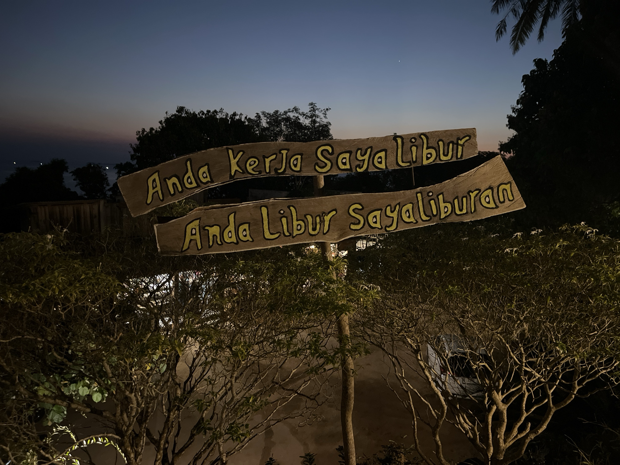 The image captures a twilight scene in Karimunjawa, Indonesia. Two weathered wooden signs, painted with yellow lettering, are prominently displayed. The top sign reads "Anda Kerja Saya Libur," and the bottom sign says "Anda Libur Sayaliburan." These phrases, when translated from Indonesian, suggest a playful message about work and vacation.
The signs are supported by a wooden post and are positioned in front of lush, dark foliage. In the background, silhouetted trees and vegetation are visible against a gradient sky that transitions from deep blue at the top to hues of orange and pink near the horizon. A faint line of lights, possibly from boats or distant buildings, is discernible on the horizon over what appears to be water. The overall atmosphere is serene and hints at an evening setting in a tropical locale.