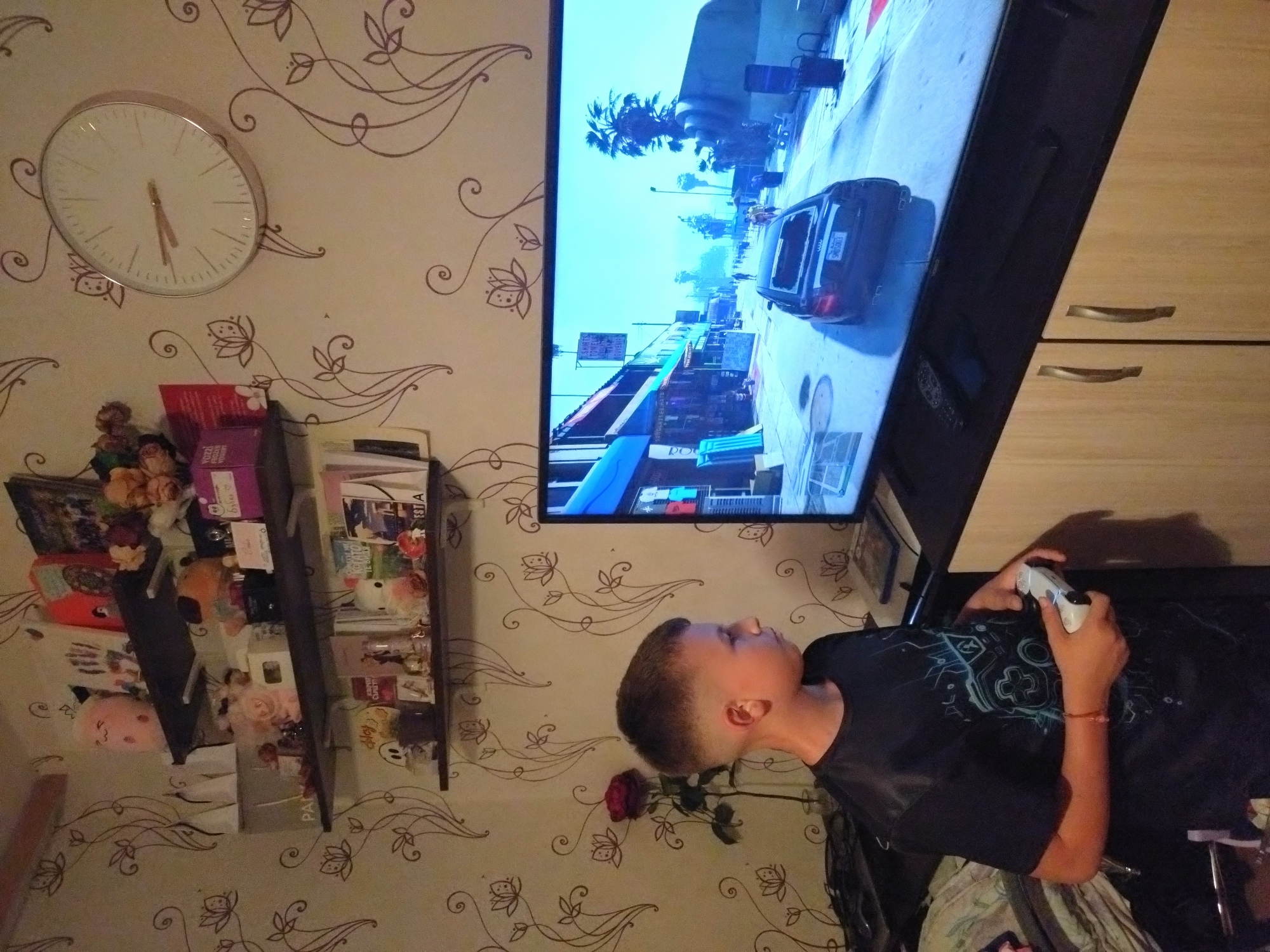 A boy is playing a video game on a television. The television is mounted on a wall next to a cabinet. The game displayed on the screen is Grand Theft Auto V, showing a street scene with a car. The boy is sitting in a chair and holding a white PlayStation controller. The wallpaper has a floral pattern. A shelf with decorations is visible to the left of the television. A clock is mounted on the wall above the shelf. The location is Stara Zagora, Bulgaria. A rose is seen under the television.