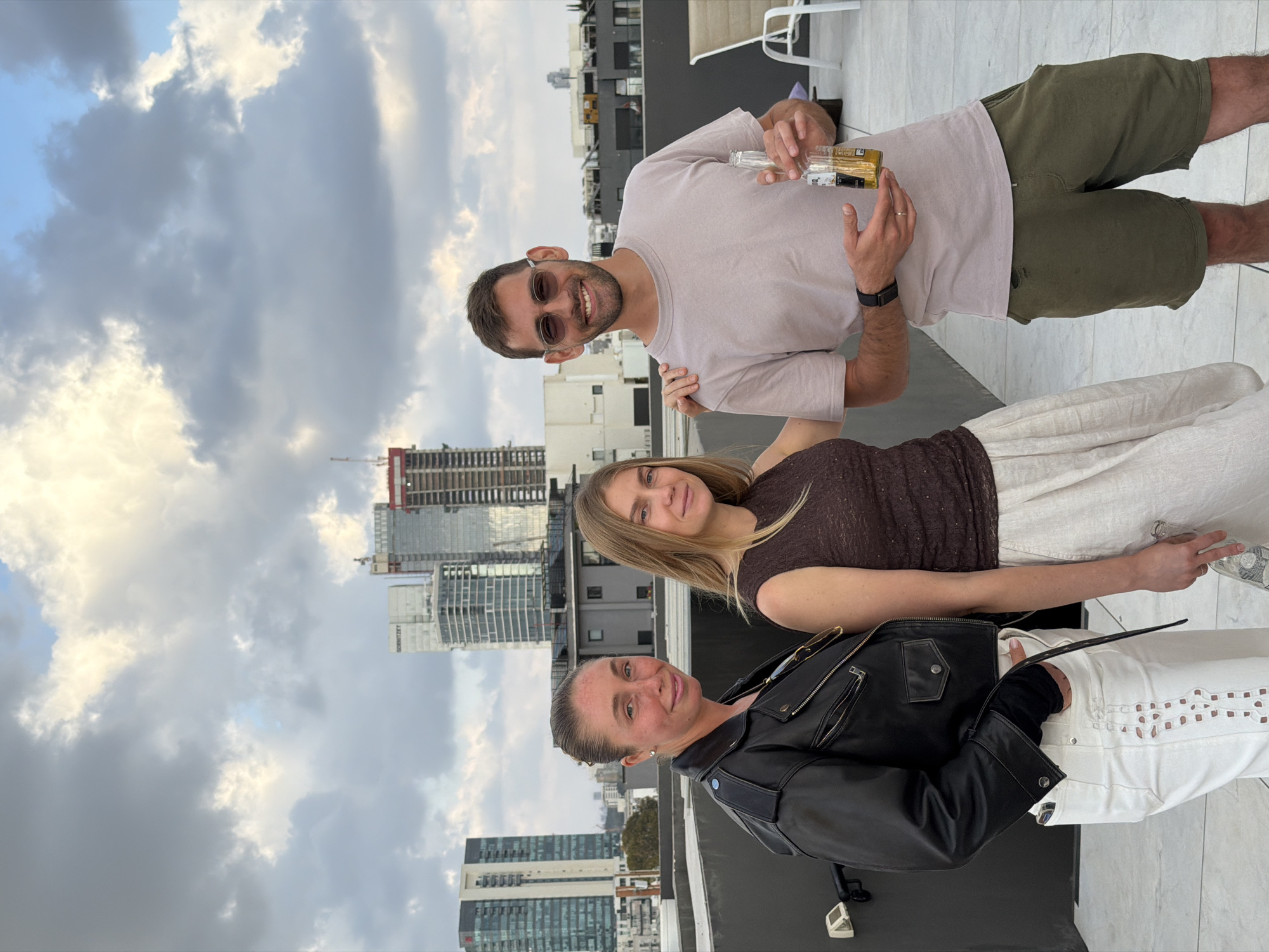 A group of three people, a man and two women, stand together on a rooftop overlooking Tel-Aviv, Israel. The man on top, seen from the waist up, is fair-skinned, wears sunglasses, a pale pink shirt, and green shorts. He holds a glass bottle in his right hand. The woman in the middle, blonde, wears a brown top and light-colored skirt. The woman on the bottom has her hair pulled back, wears a black leather jacket and white pants. Behind them is the cityscape of Tel-Aviv, including modern high-rise buildings, under a cloudy sky.
