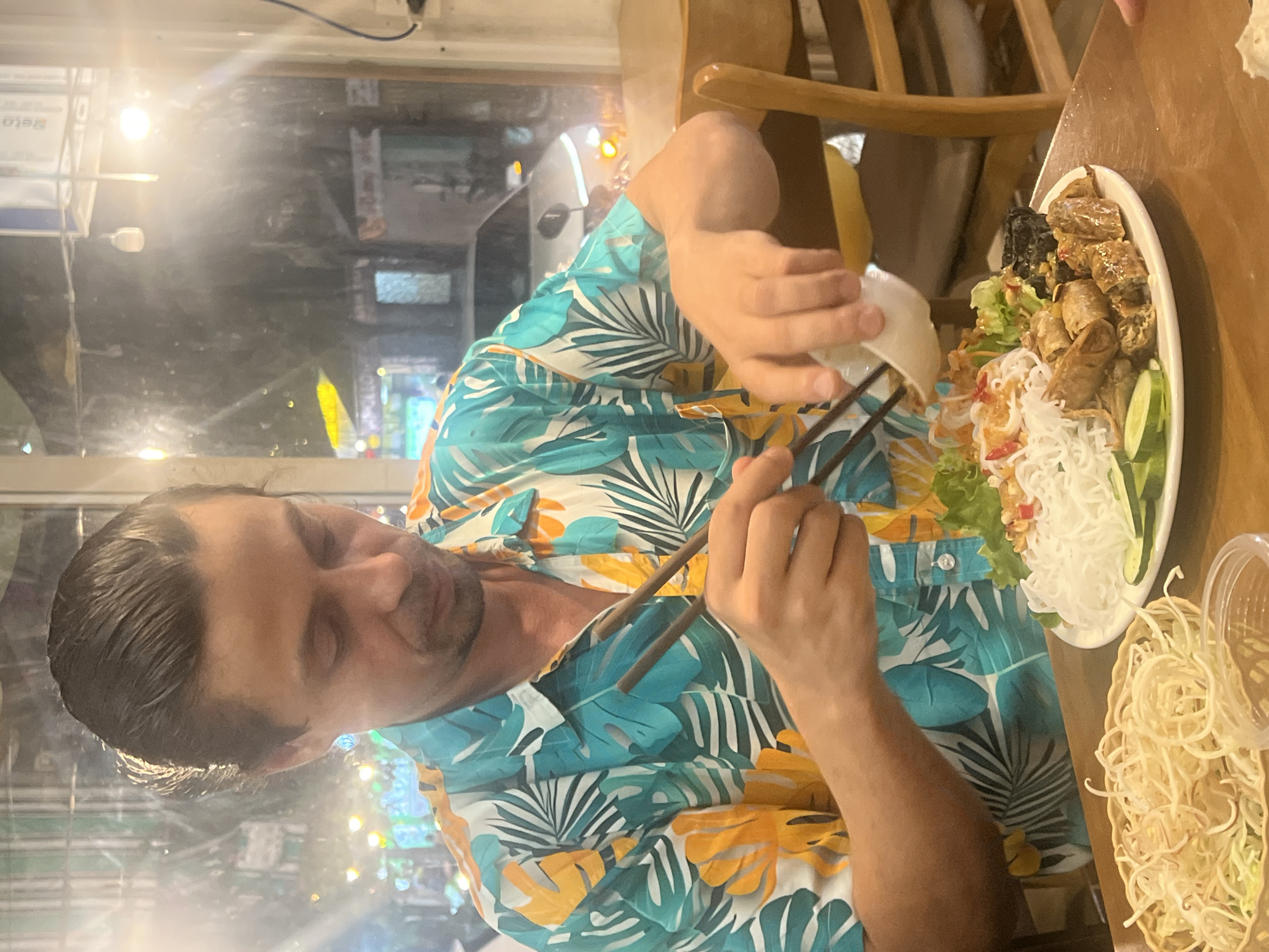 A man with dark hair pulled back is seated at a table in a restaurant in Da Nang, Vietnam, wearing a tropical-print shirt. He is actively eating a meal with chopsticks, holding a small white bowl in his left hand and a portion of food with his chopsticks in his right. In front of him is a plate containing rice noodles, lettuce, spring rolls, and cucumber slices. To his left, another basket holds shredded vegetables or noodles. The restaurant interior is dimly lit with reflections visible in a large window behind him, showing a street scene with lights and a car. The overall setting suggests an indoor dining experience.