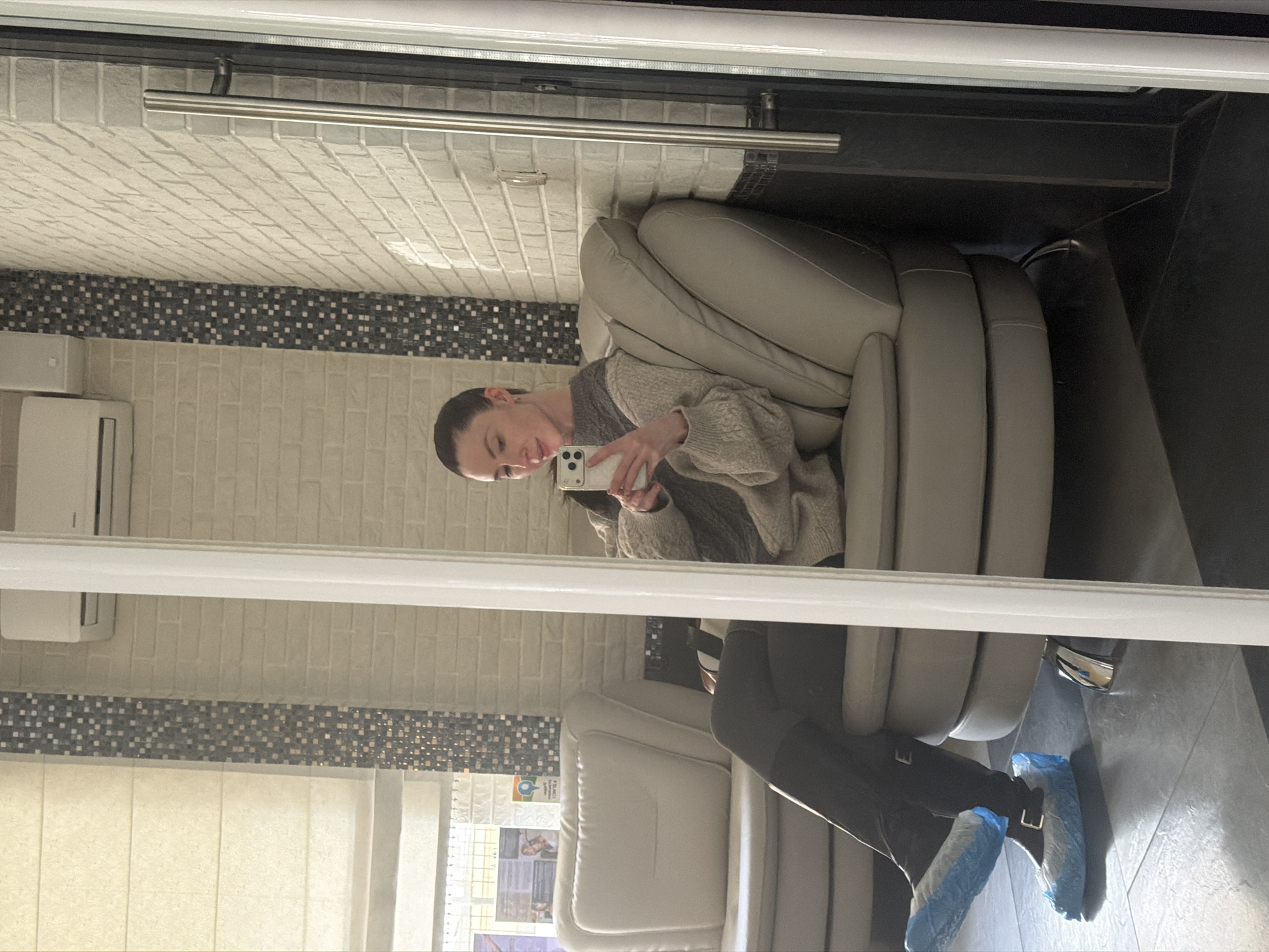 The image, taken in Perm, Russia, depicts a mirror reflection of a woman sitting comfortably in a light grey, padded chair, actively taking a selfie with her white smartphone. She is wearing a light-colored, textured sweater and dark trousers, with her dark hair pulled back. Her feet are visible, covered in blue disposable shoe covers, indicating she is in an environment where cleanliness is prioritized, such as a medical office or a salon.
The indoor setting features walls adorned with light beige brick-patterned tiles, accented by a decorative horizontal band of small black and white mosaic tiles. A white air conditioning unit is mounted on one wall. The floor is covered in dark grey tiles. In the background, light-colored vertical window blinds are visible, suggesting it is daytime. A metal bar or handle is seen near the top of the mirror's frame. Reflected faintly on a wall behind the woman is a display or poster, but any text is too indistinct to be legible. The woman's expression is neutral as she focuses on her phone. The overall scene conveys a clean, modern, and possibly professional environment.