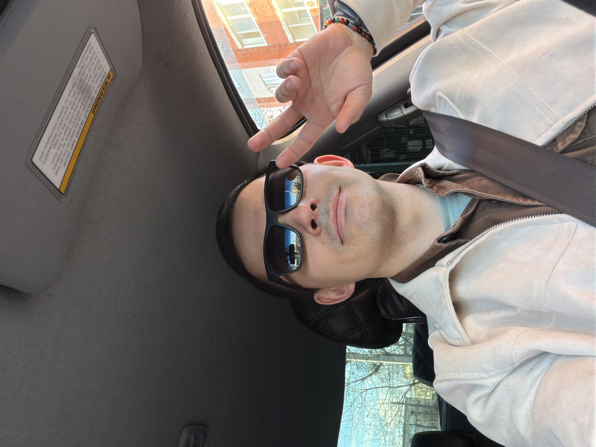 This image is oriented 90 degrees counter-clockwise, depicting a man taking a selfie from inside a car during the daytime. The man, positioned in the foreground, is wearing dark rectangular sunglasses, a light cream-colored hooded jacket over a brown and light blue inner layer, and a multi-colored beaded bracelet on his right wrist. He has a subtle smile and holds his right hand up in a "peace" or "victory" gesture.
He appears to be seated in the front passenger or driver's seat, with a dark grey seatbelt crossing his chest. Part of a dark, possibly leather, quilted car seat headrest is visible behind his right ear. The car's interior features a grey headliner and a sun visor.
Through the car windows, an urban scene is visible. A multi-story brick building with numerous windows occupies the upper part of the background. Below this, bare tree branches are silhouetted against a clear blue sky, visible directly and as a reflection in the man's sunglasses. The overall mood appears casual and relaxed.
A prominent yellow warning label on the sun visor contains safety information, stating: "WARNING DEATH or SERIOUS INJURY can occur. Children 12 and under can be killed by the air bag. The BACK SEAT is the SAFEST place for children. NEVER put a rear-facing child seat in the front. Sit as far back as possible from the air bag. ALWAYS Use SEAT BELTS and CHILD RESTRAINTS. See the Owner's Manual for further information and explanations." The location context provided for this image is Izhevsk, Russia.