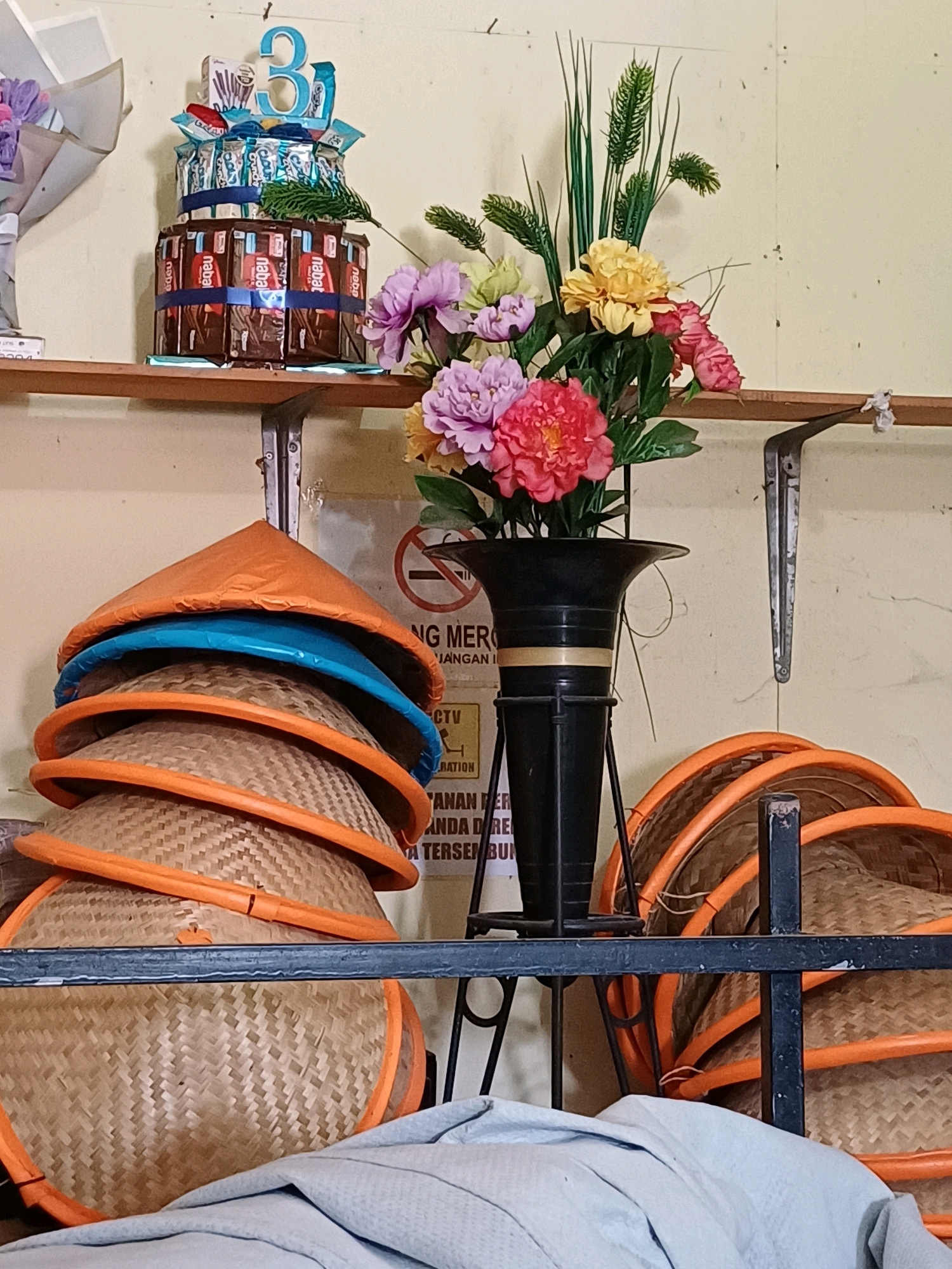 A two-tiered chocolate bar cake with a blue "3" topper sits on a shelf, accompanied by a bouquet of artificial flowers in a black vase. Below, a stack of conical hats, some orange and blue, are arranged next to more hats on a metal rack. A sign with a "no smoking" symbol and text in Indonesian is visible on the wall behind the hats. The scene appears to be indoors, possibly a shop or storage area, given the shelving and wall material.
