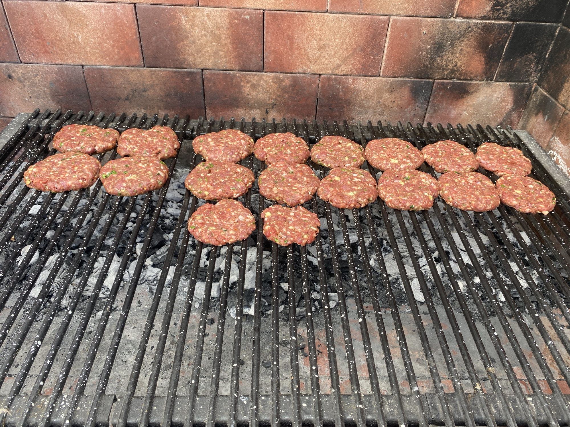 The image shows a grill filled with approximately 18 uncooked meat patties arranged in a somewhat organized grid pattern. The grill is positioned over hot coals, visible beneath the metal bars. The backdrop features a brick wall with a reddish-brown hue, possibly part of an outdoor cooking area. The patties appear to be seasoned with herbs or spices. The scene depicts an outdoor cooking activity. There is no information available in the image about the location being in Buenos Aires, Argentina.